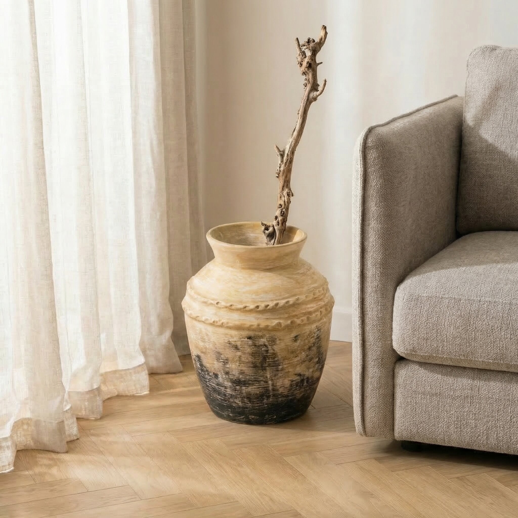 Beige vase with a branch on a wooden floor next to a beige sofa.