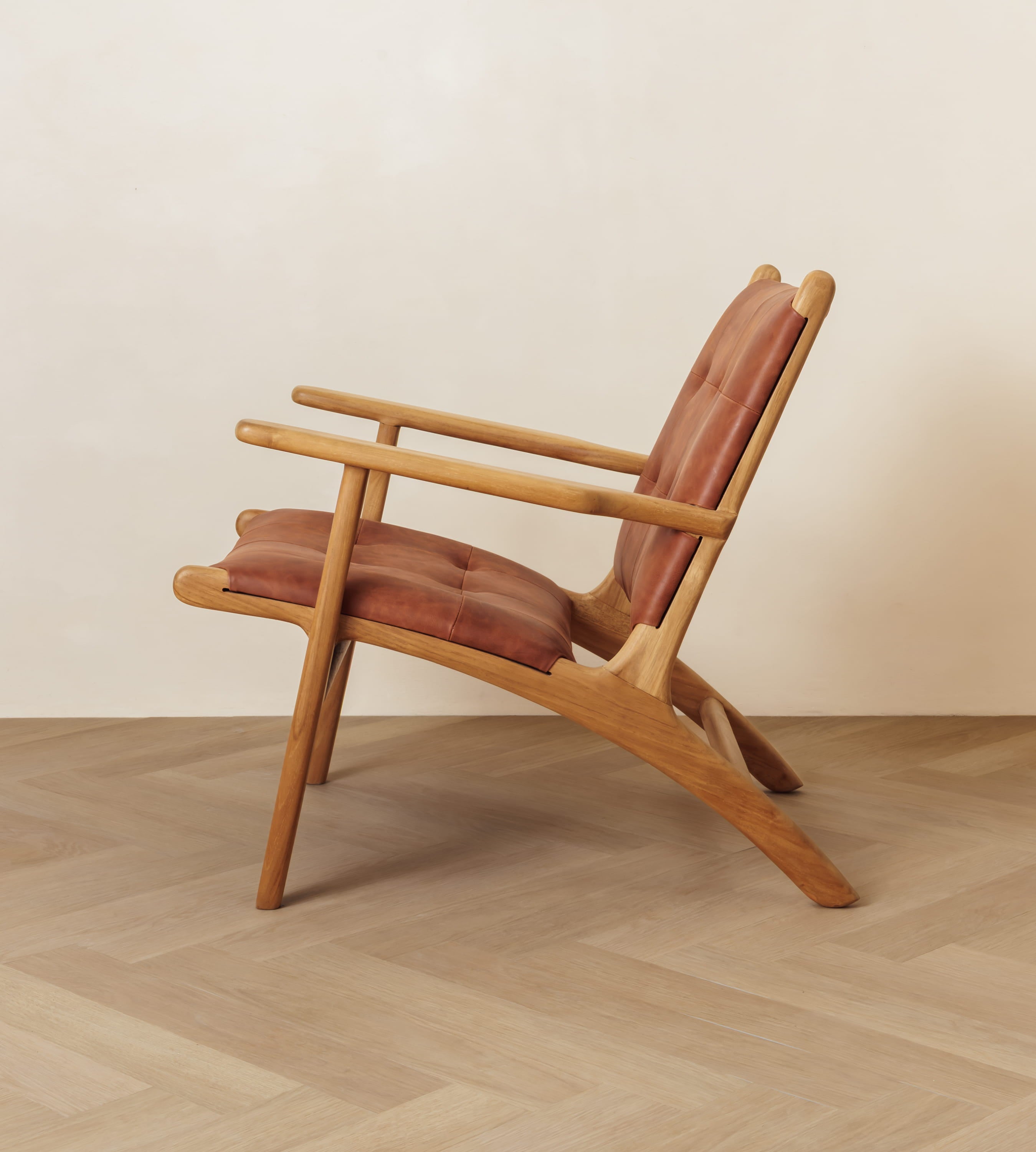 A mid-century modern wooden lounge chair with reddish-brown upholstered seat and back cushions, shown on a light wood herringbone floor against a plain beige wall.