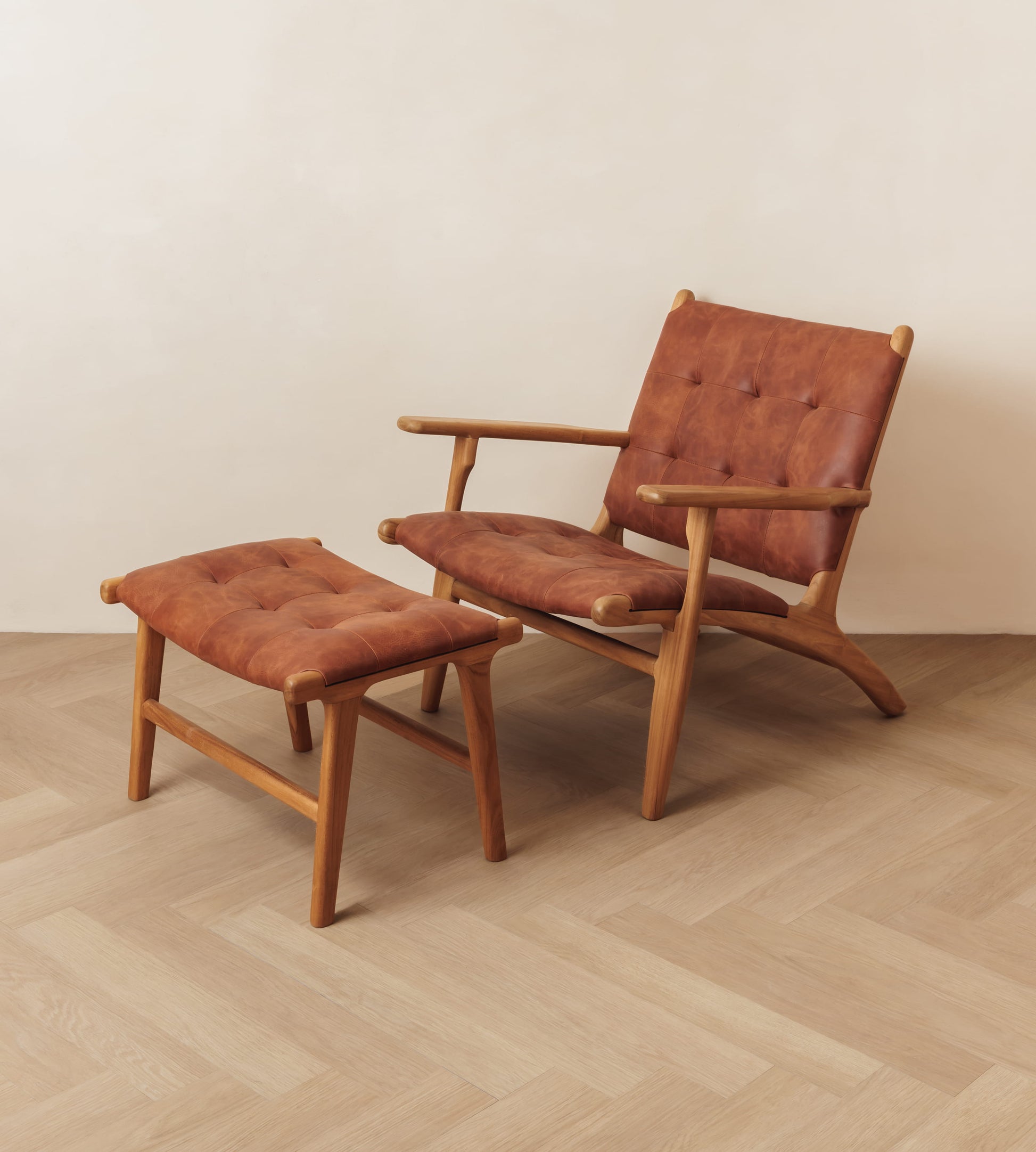 A mid-century modern wooden lounge chair with tufted brown leather upholstery, shown with a matching ottoman on a light wood herringbone floor against a plain beige wall.