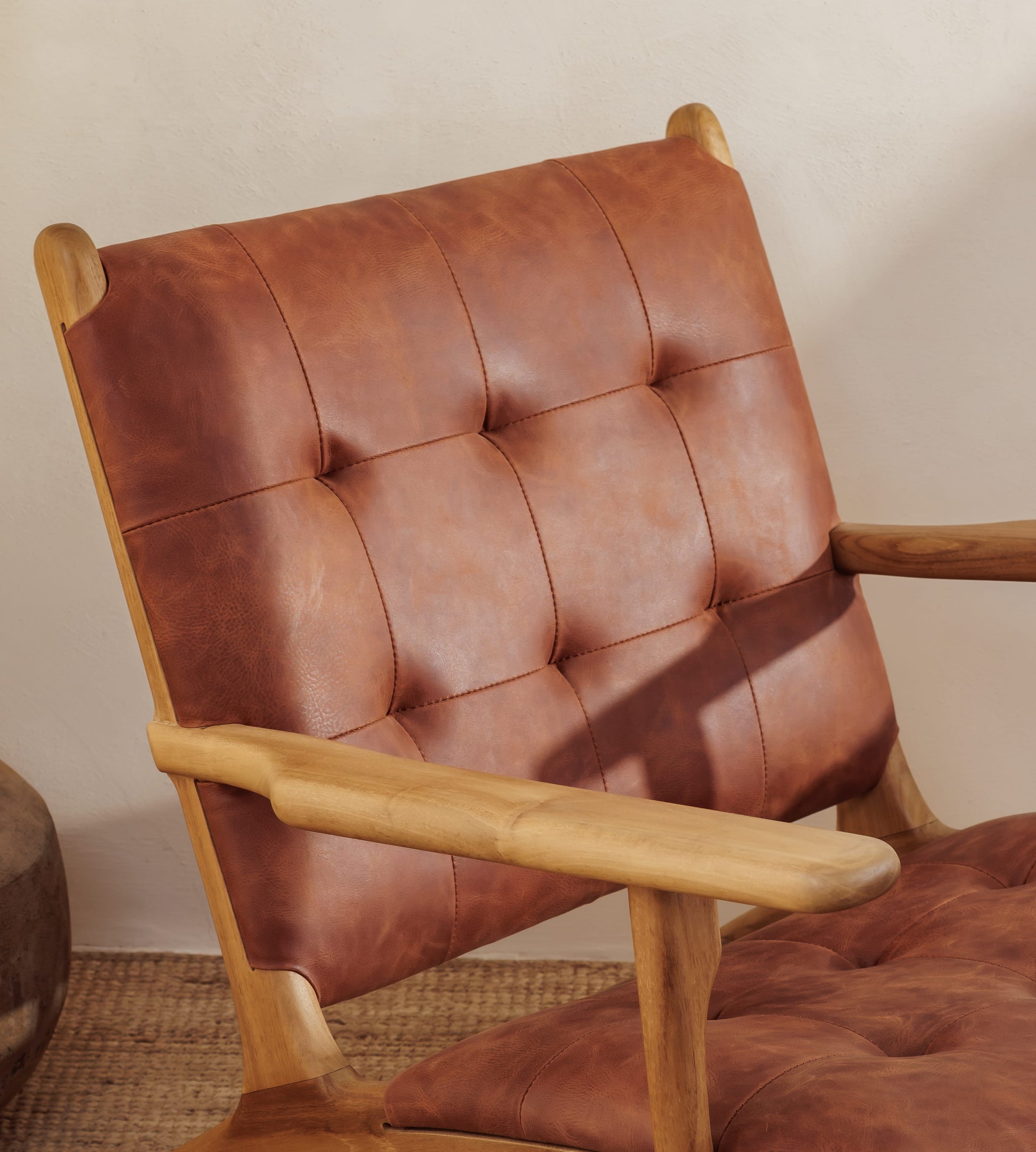 A close-up of a a modern leather armchair with a tufted brown leather backrest and seat cushion, set against a neutral beige wall.