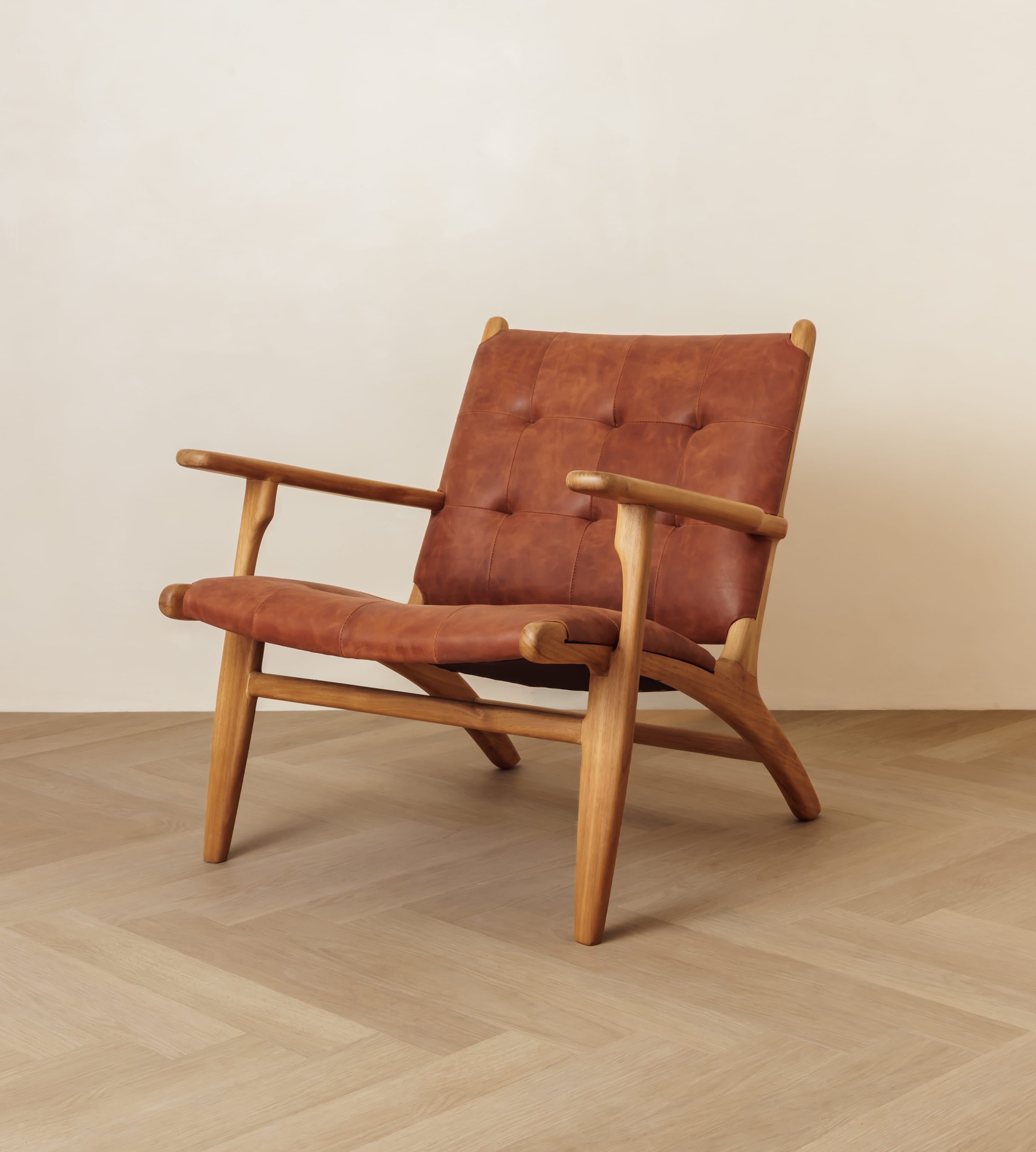 A mid-century modern wooden armchair with a tufted reddish-brown leather seat and backrest, set against a minimalist beige wall and light wood herringbone floor.