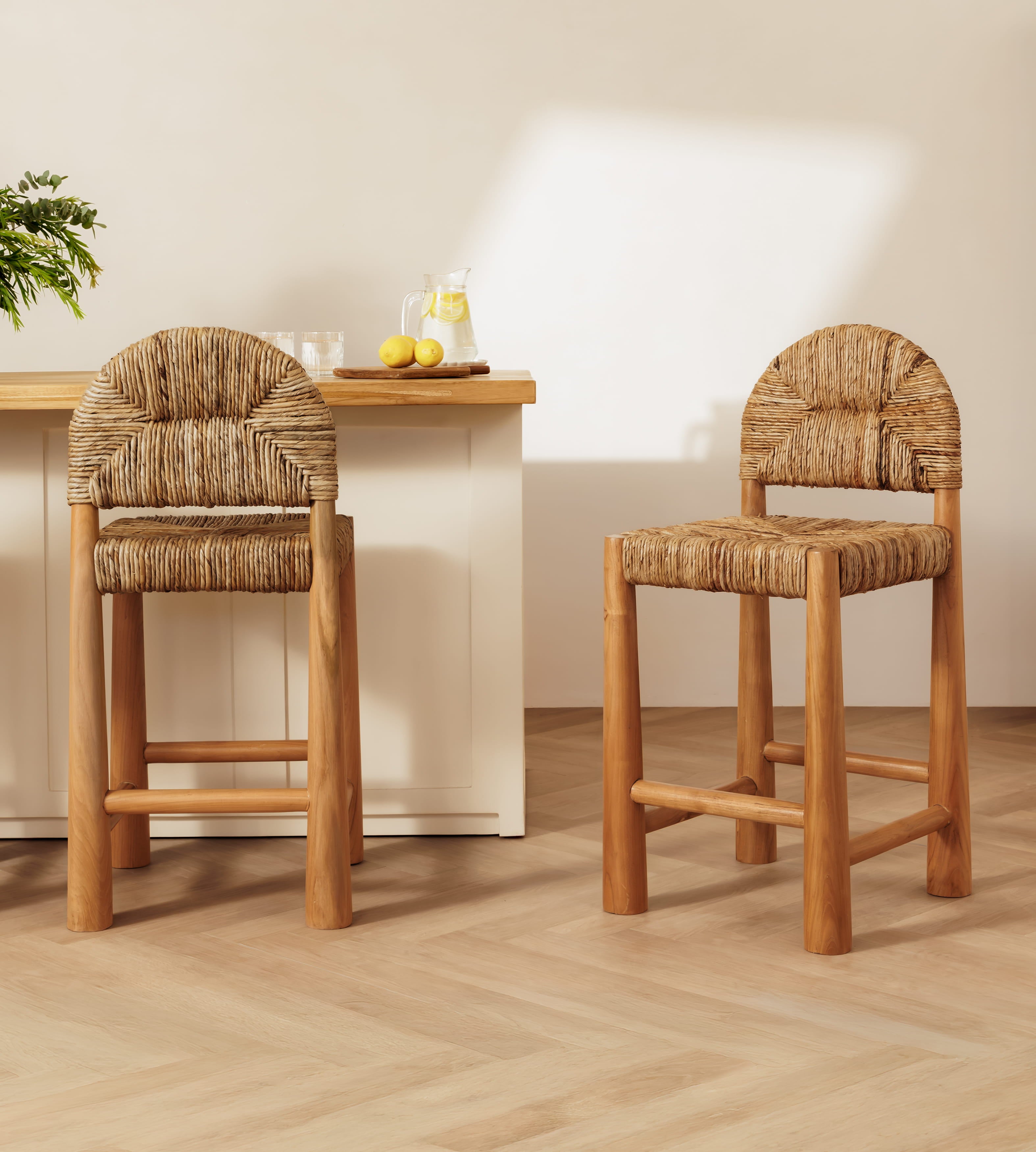 Two banana leaf bar stools with woven straw seats and backrests, shown in a bright, modern kitchen setting with a light wood island and pale walls.
