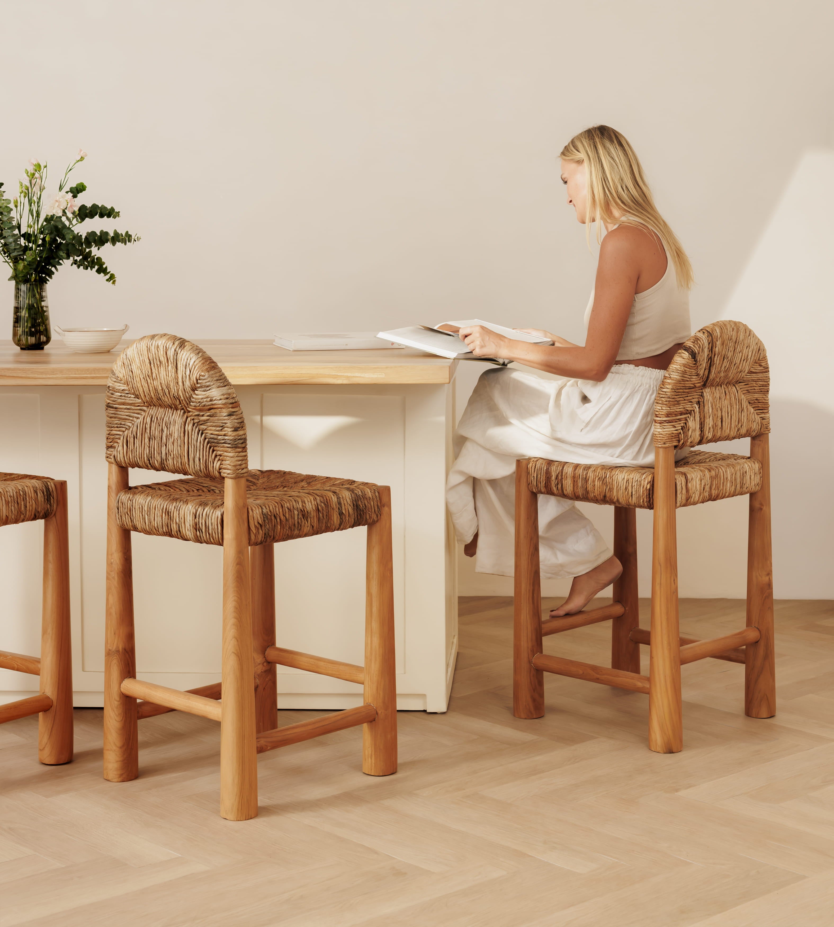 A woman sits at a tall banana leaf bar stool beside a light wood counter in a bright, minimalist interior. Two matching bar stools are visible, and a vase with greenery sits on the counter.