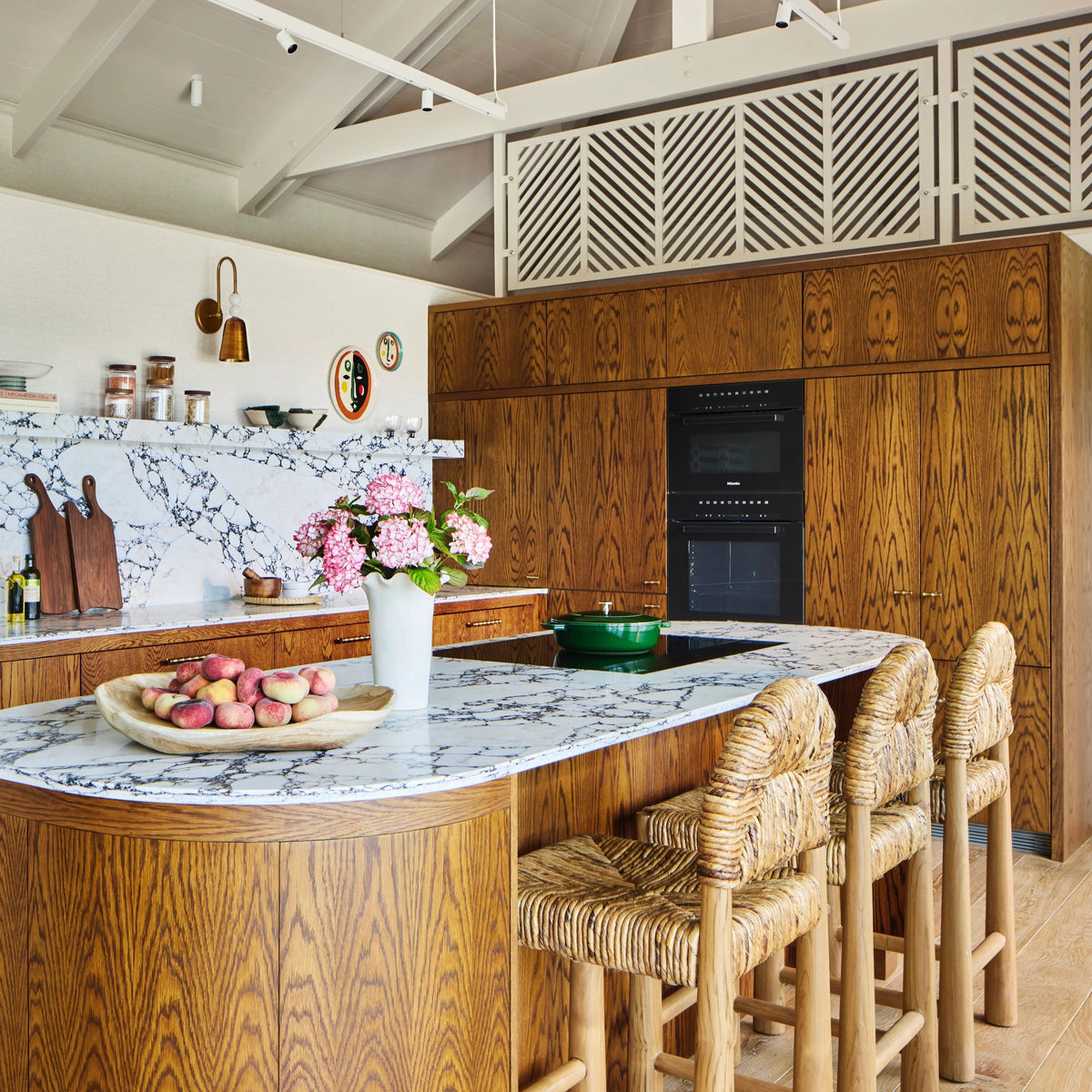 A modern kitchen interior with warm wood cabinetry and island, a white marble backsplash, woven bar stools, and a countertop bowl of fruit. No person or animal is present, but this is still a real interior environment rather than a clean isolated product shot, so subject_type is Lifestyle.