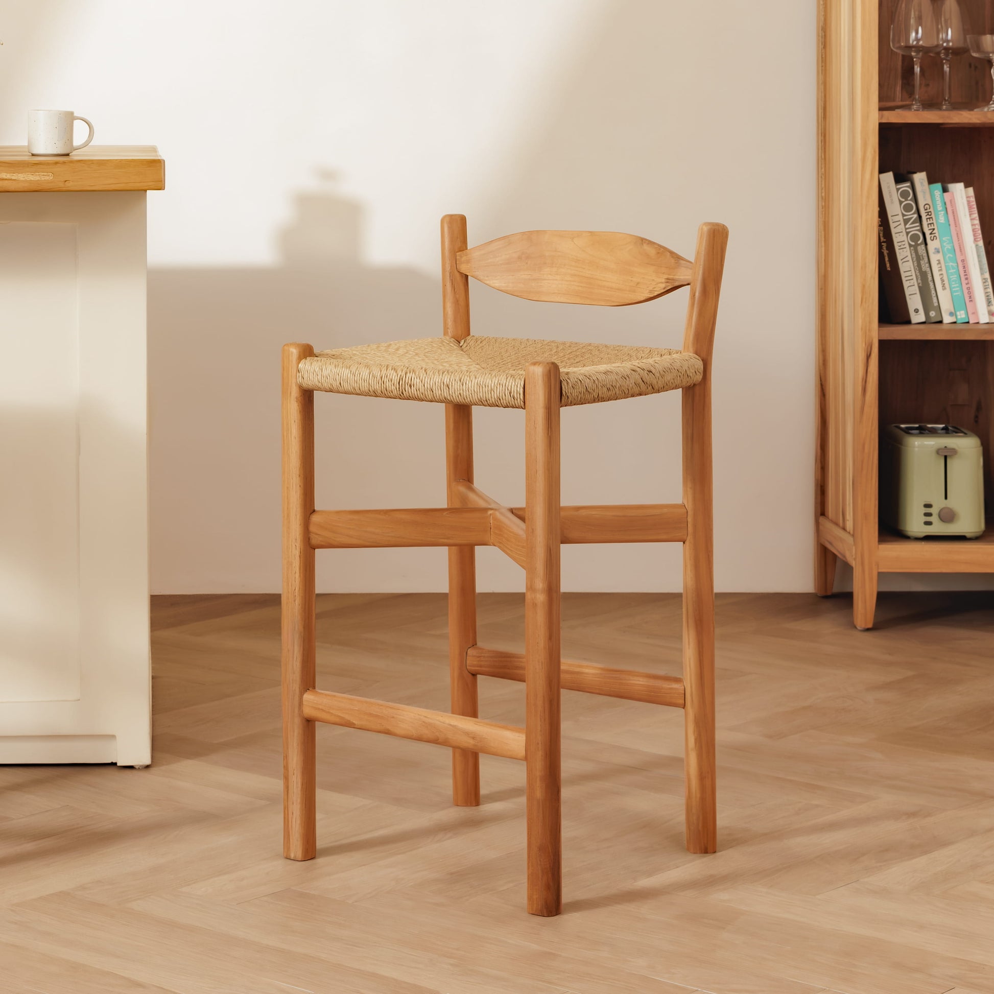 A wooden bar stool with a woven straw seat and curved wooden backrest, shown in a bright, modern room with a light wood floor, a white side table, and a wooden bookshelf.
