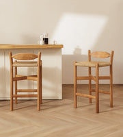 Two wooden bar stools with woven seats are shown in a bright, minimalist interior with a light wood counter and a cream wall.
