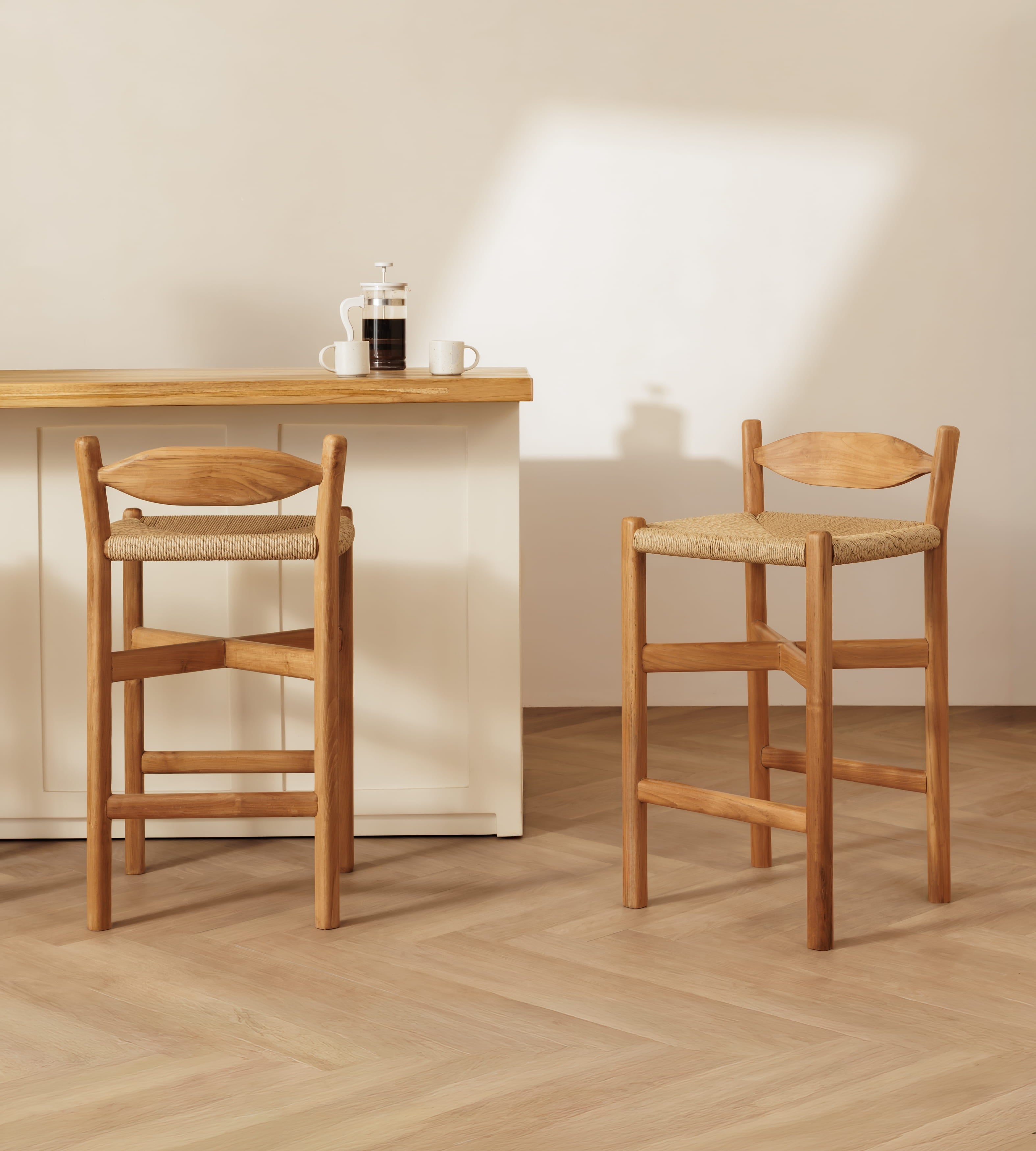 Two wooden bar stools with woven seats are shown in a bright, minimalist interior with a light wood counter and a cream wall.