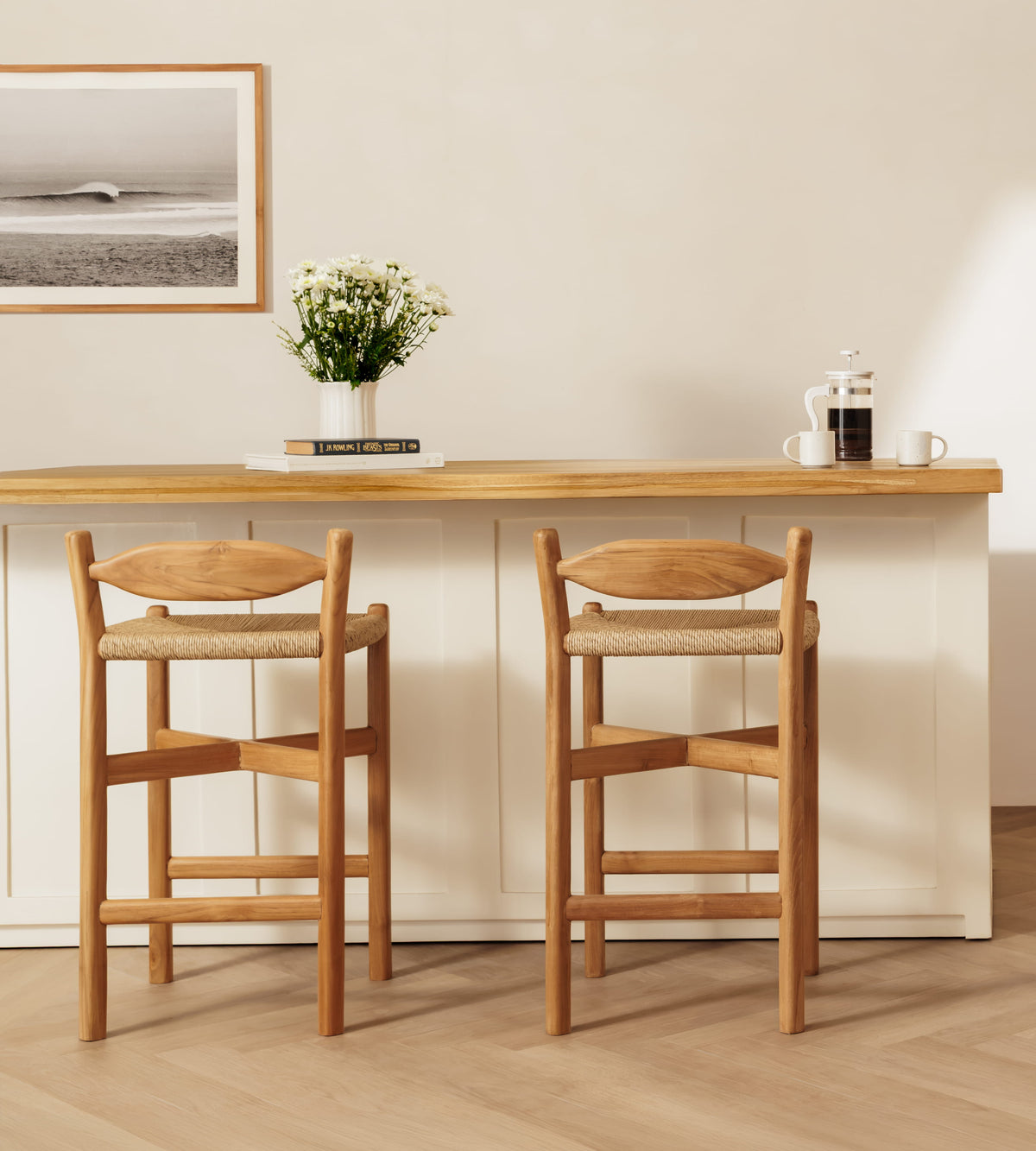 Two wooden bar stools with woven seat cushions positioned in front of a matching wooden-topped counter in a bright, minimalist interior.
