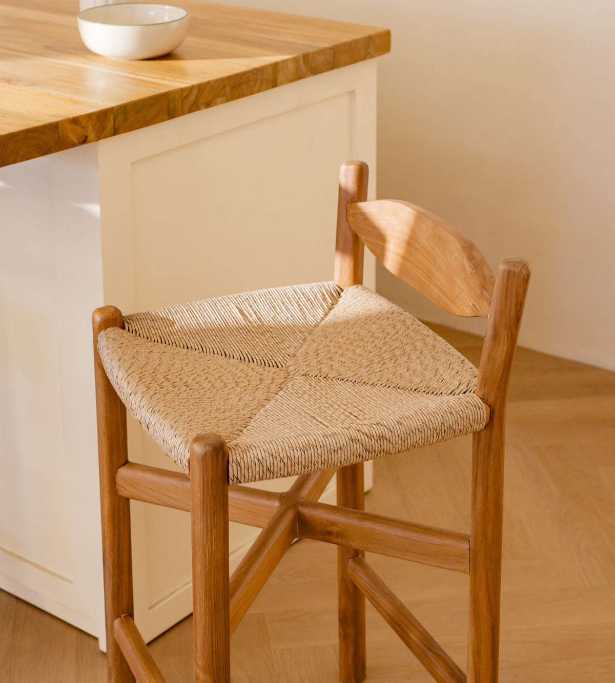 A wooden bar stool with a woven rope seat and backrest, shown in a bright kitchen setting next to a wooden countertop.