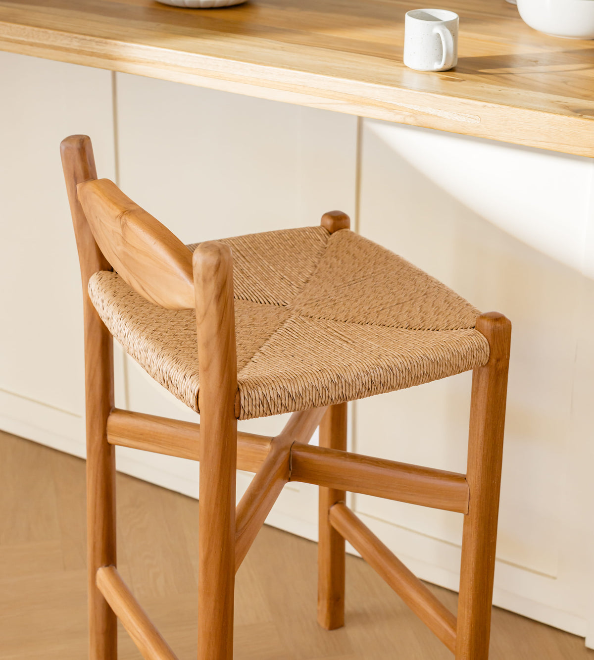 A wooden bar stool with a woven rope seat, shown in a bright kitchen setting with a wooden countertop and white cabinets in the background.