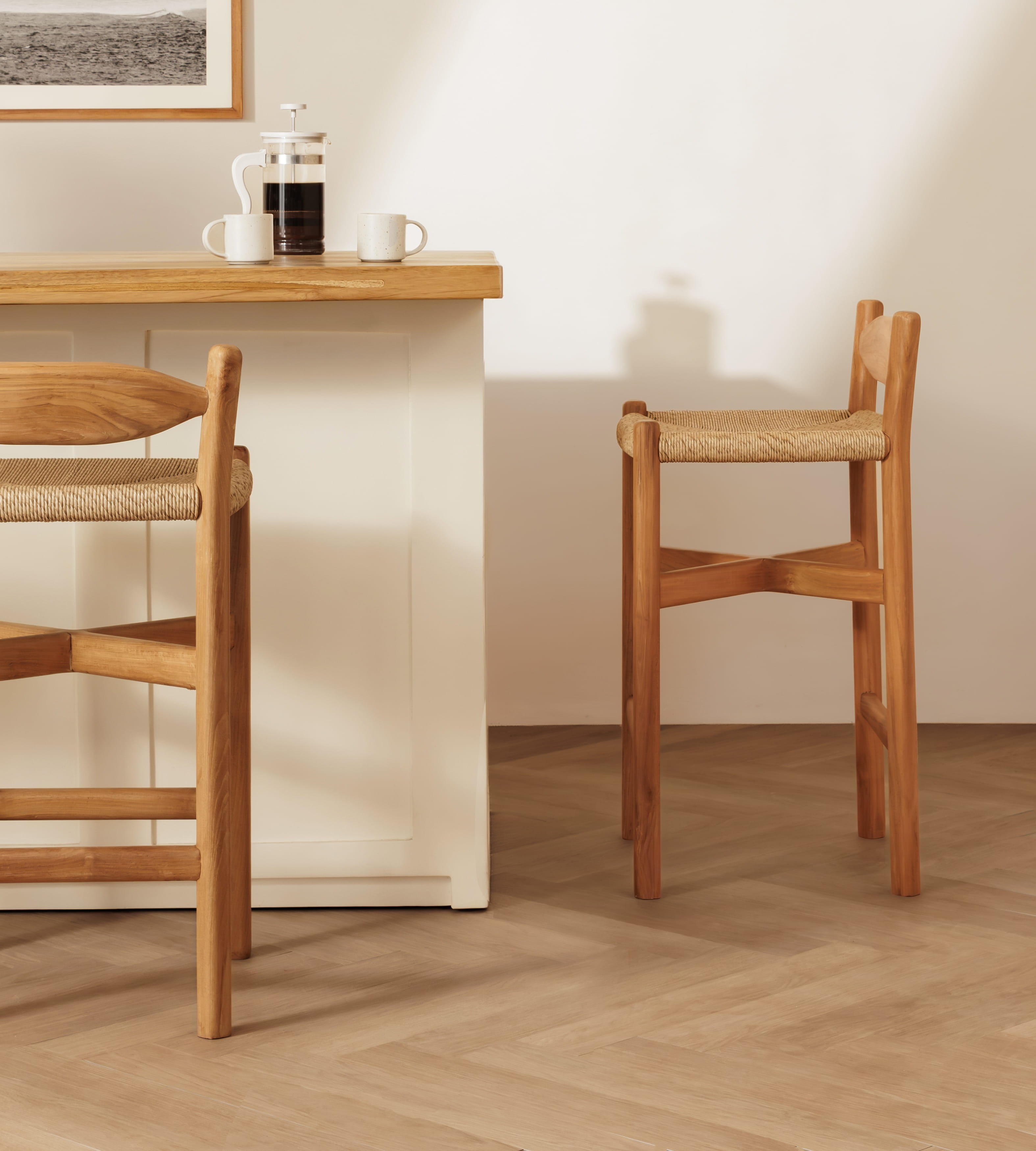 A warm-toned wooden bar stool with a woven seat cushion stands beside a light wood and white counter in a minimalist interior setting.