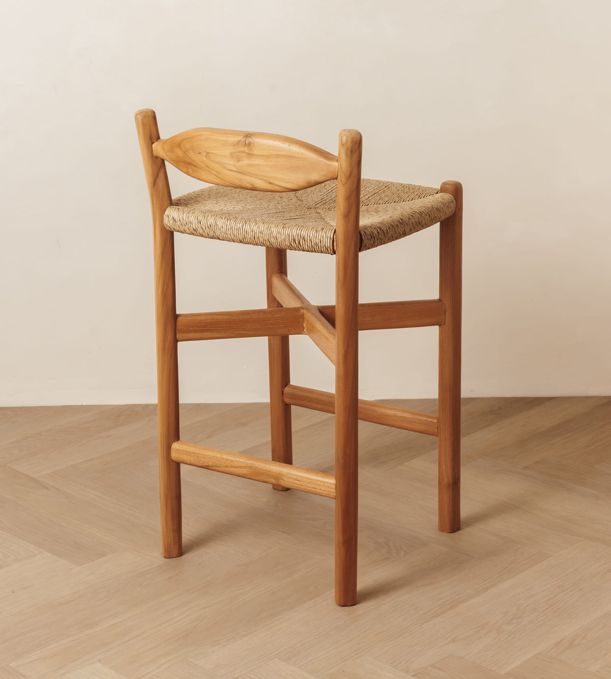 A wooden bar stool with a woven rope seat and a curved wooden backrest, shown in a minimalist room setting with a light beige wall and herringbone wood floor.