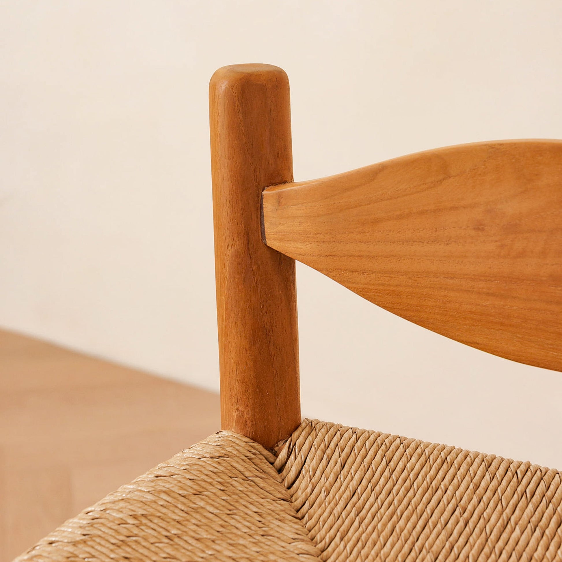 Close-up of a wooden chair corner with a warm medium-brown wood frame and a woven natural-fiber seat against a light beige wall.