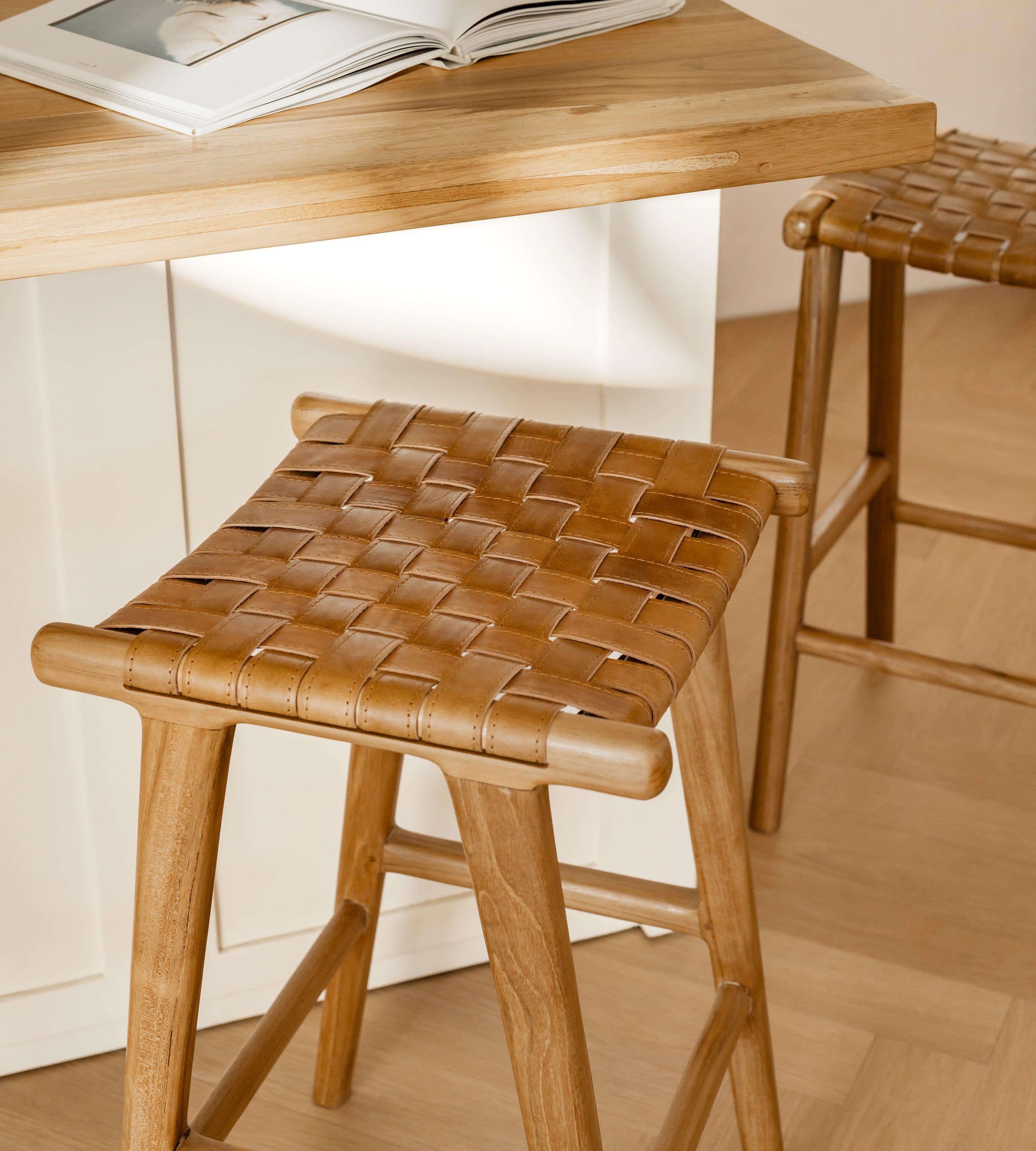 A wooden bar stool with a woven leather seat is shown in a kitchen setting next to a wooden counter, with another matching stool partially visible in the background.