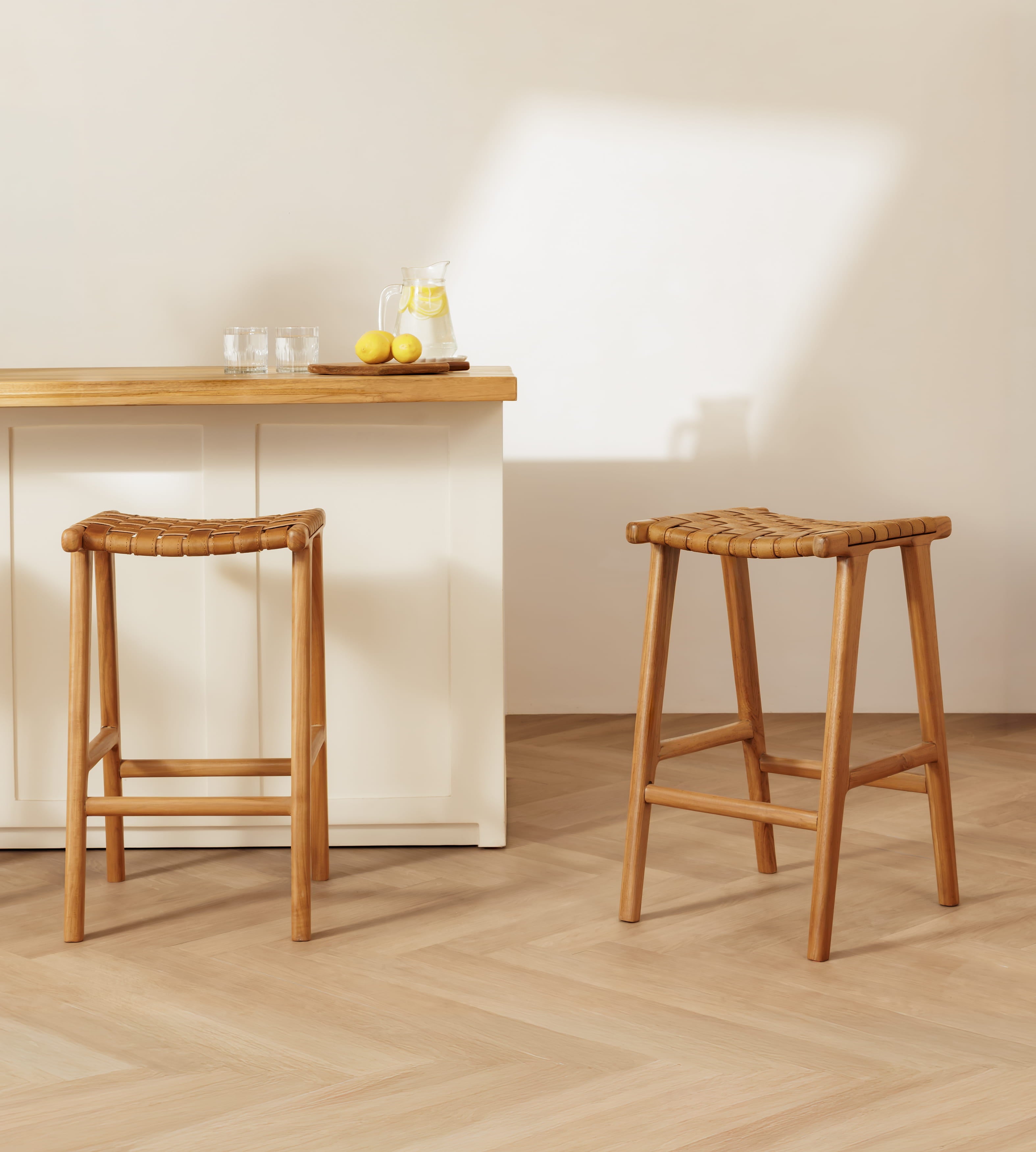 Two leather bar stools with woven seats are shown in a bright, modern kitchen setting with a light wood island and a cream wall.