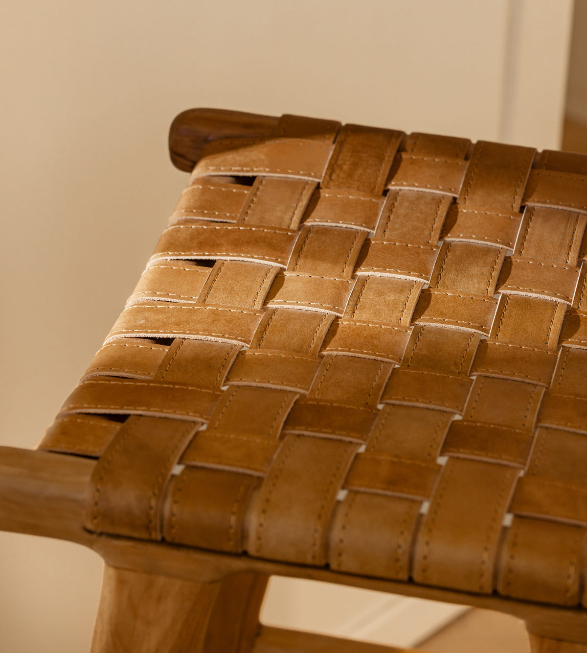 Close-up of a woven leather seat strap stool with a warm tan-brown leather seat and wooden frame, set against a neutral beige background.