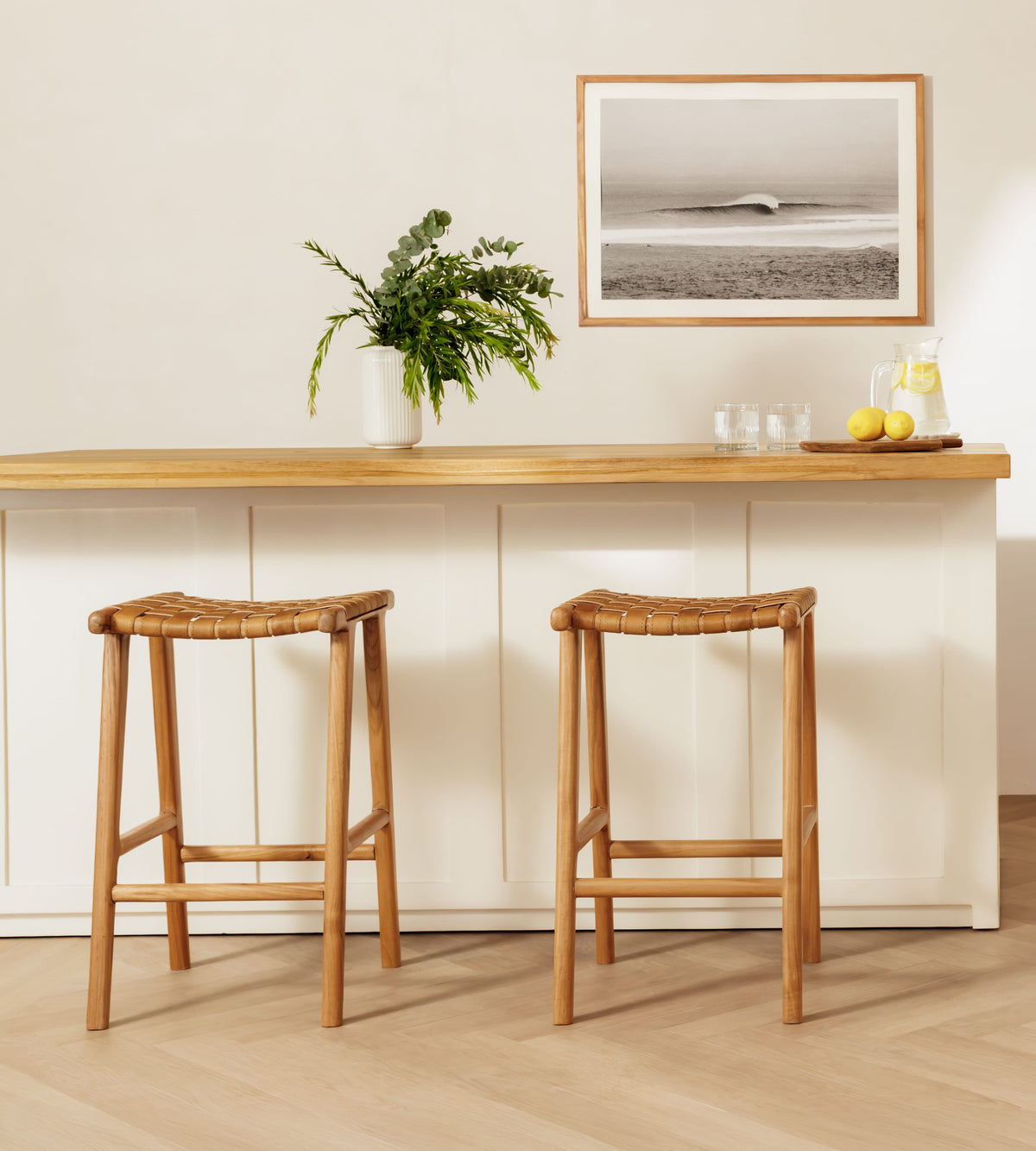 Two wooden bar stools with woven seats placed in front of a white kitchen island with a light wood countertop. A framed black-and-white beach print hangs on the wall above, with a potted plant and small table decor on the counter.