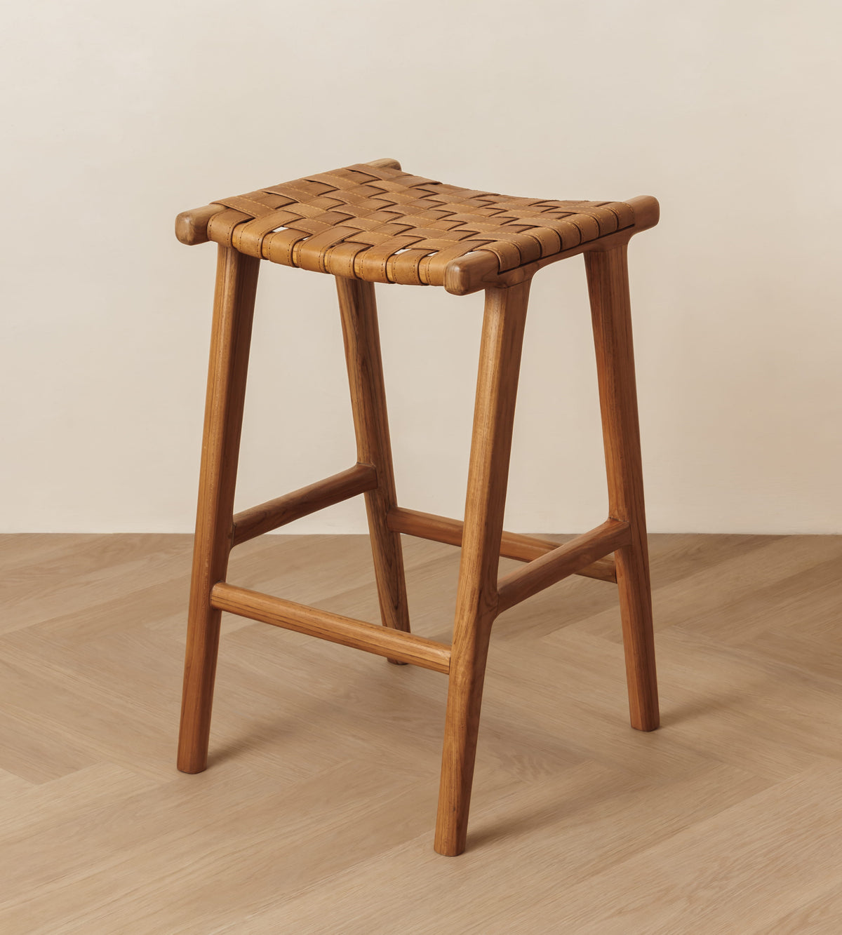 A wooden bar stool with a woven seat, shown in a minimalist room setting with a light beige wall and herringbone wood floor.
