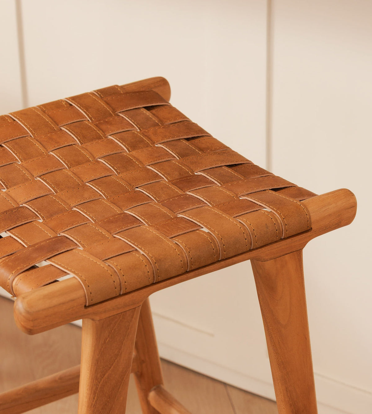 A close-up of a wooden stool with a woven tan leather seat, set against a neutral cream-colored paneled wall and light wood floor.
