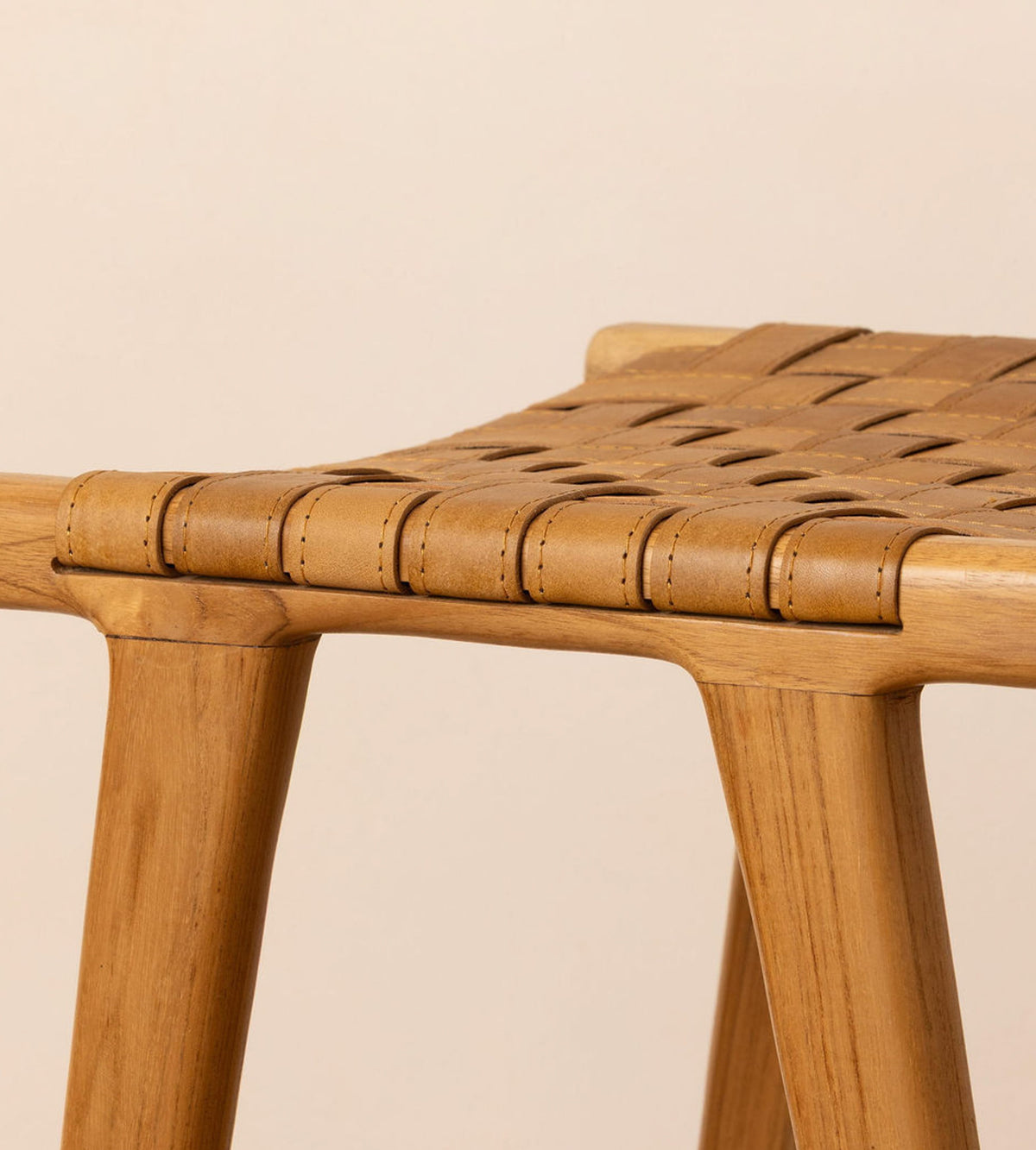 Close-up of a wooden stool with a woven seat strap in a warm tan color, set against a solid light beige background.