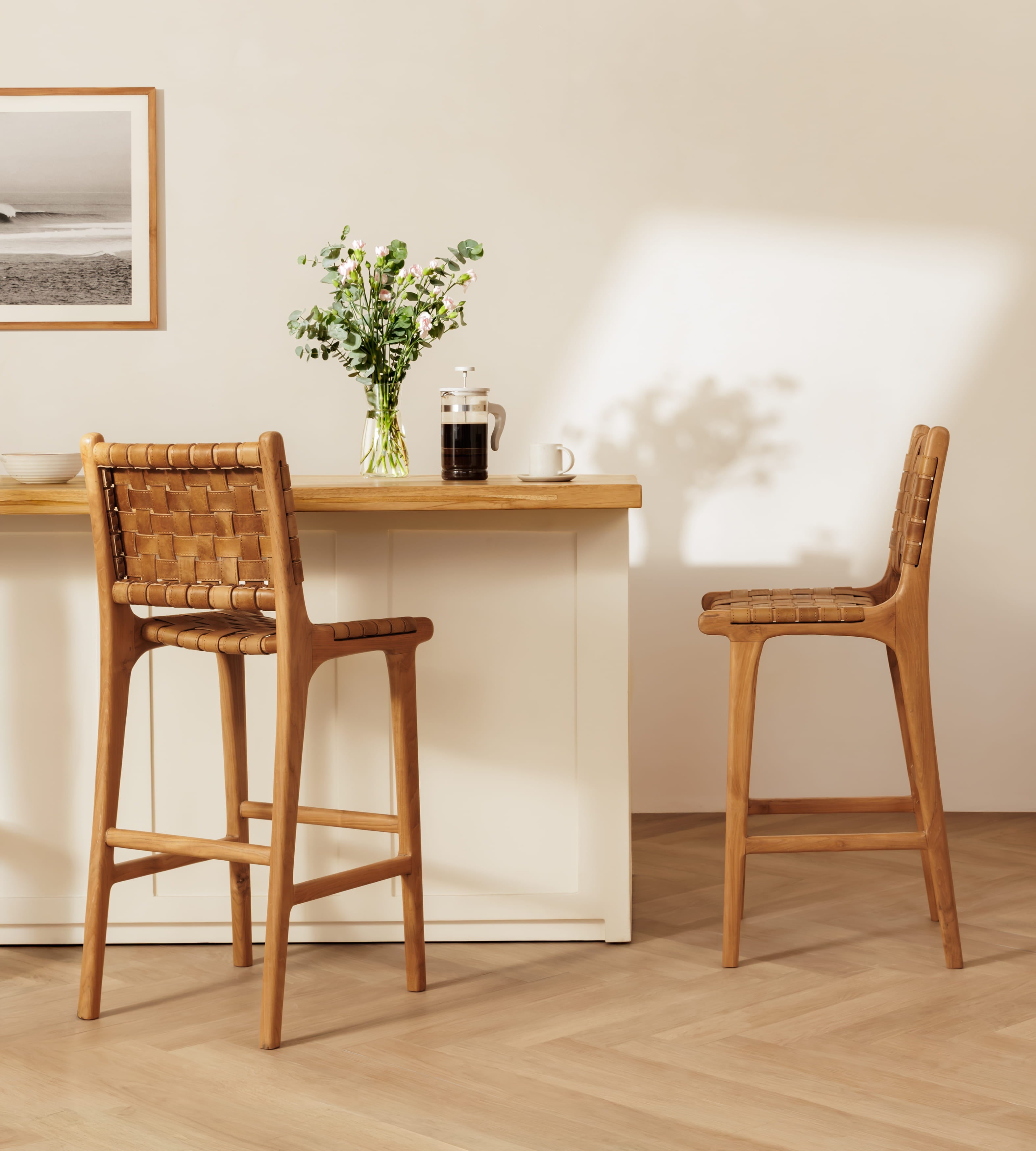 Two leather bar stools with woven seats and backs are shown in a bright, modern interior setting with a wooden counter, a vase of flowers, and warm sunlight casting shadows on the wall.