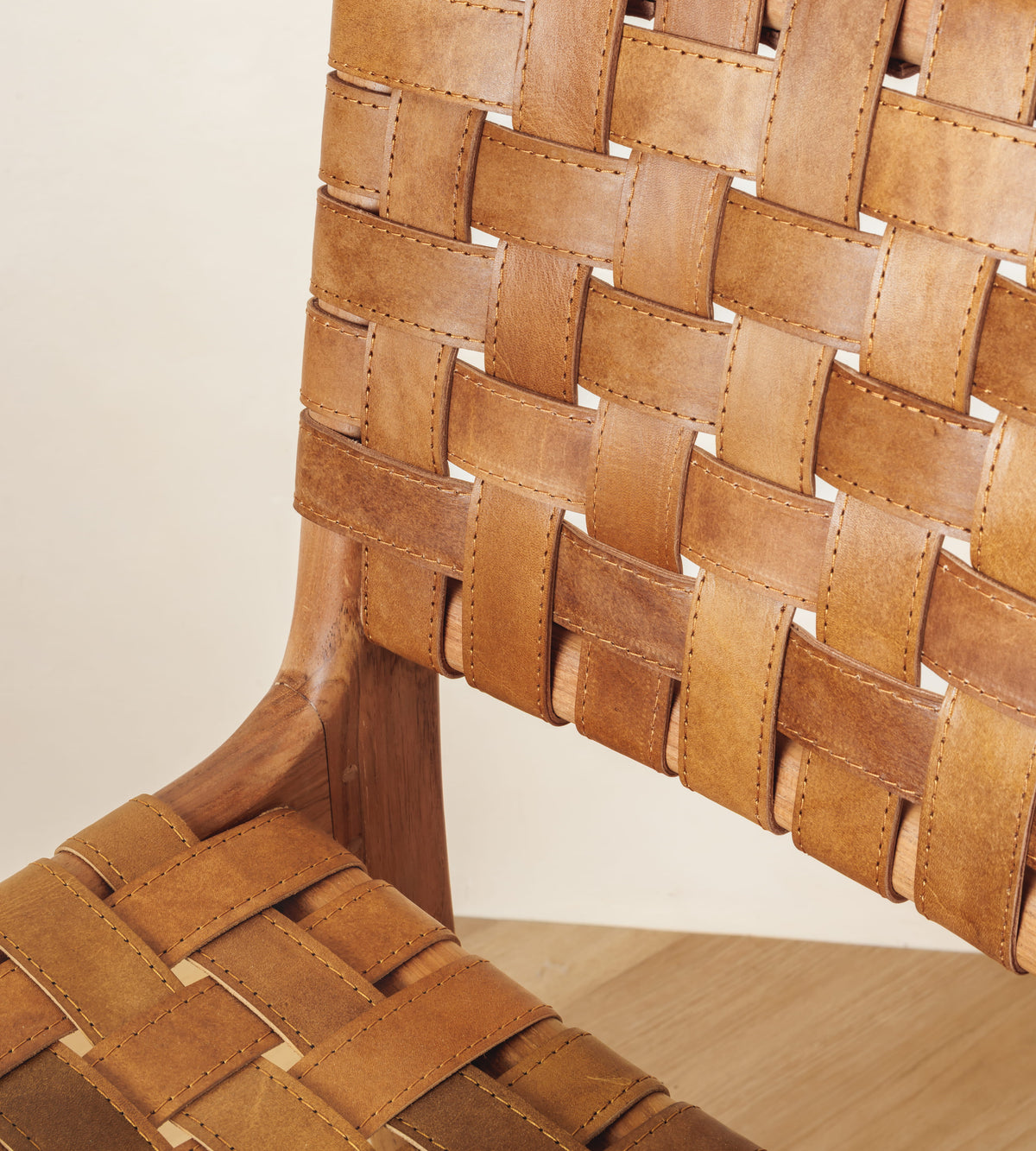 Close-up of a woven leather chair back and seat in warm tan and brown tones with a wooden frame, set against a light neutral wall and pale wood floor.