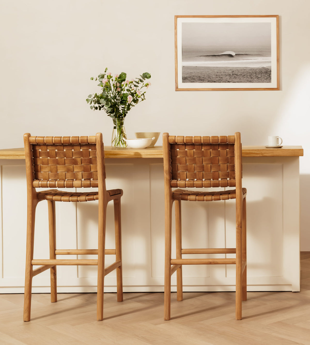 Two wooden bar stools with woven leather backs and seats, positioned in front of a light wood counter in a bright, modern kitchen setting.