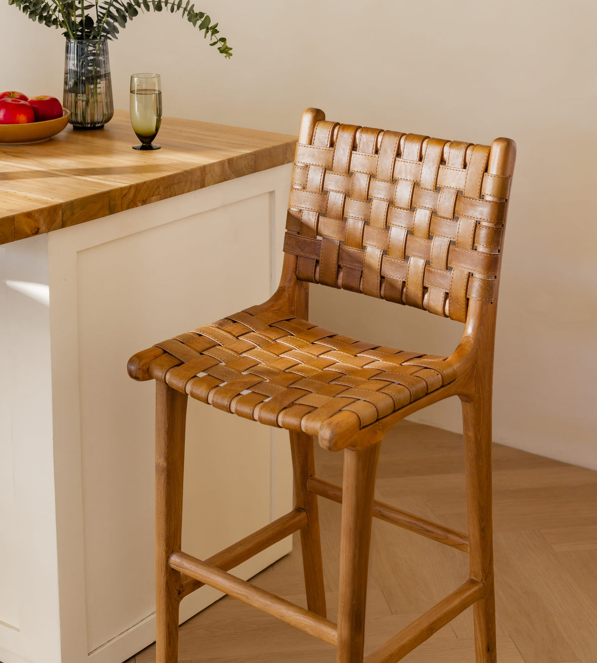 A wooden bar stool with a woven leather seat and backrest, positioned next to a kitchen island with a wood countertop and white cabinets.