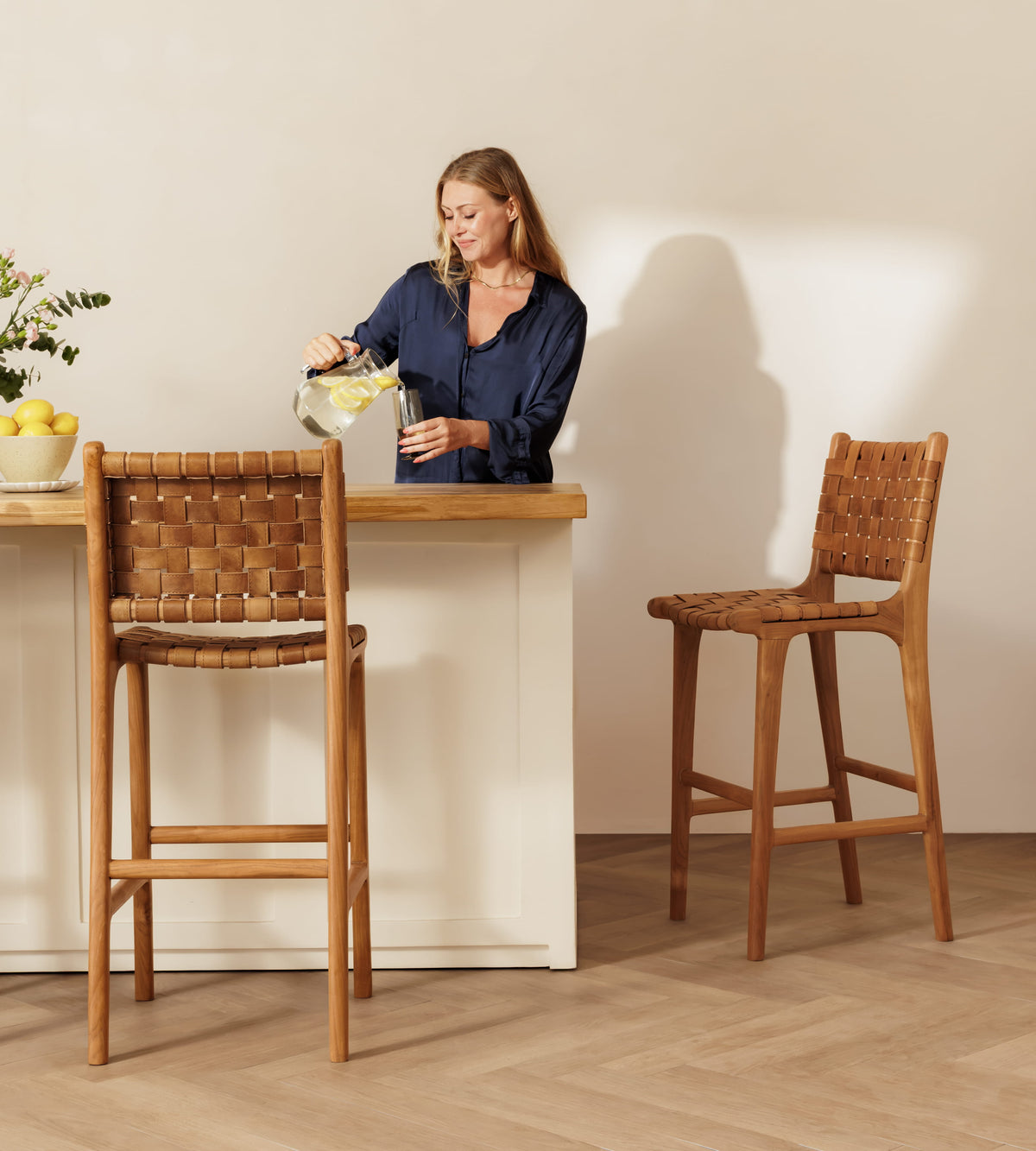 A woman stands at a light wood bar counter pouring a drink, with two woven-back bar stools in a warm neutral interior. One stool is in front of the counter and one is placed to the right.