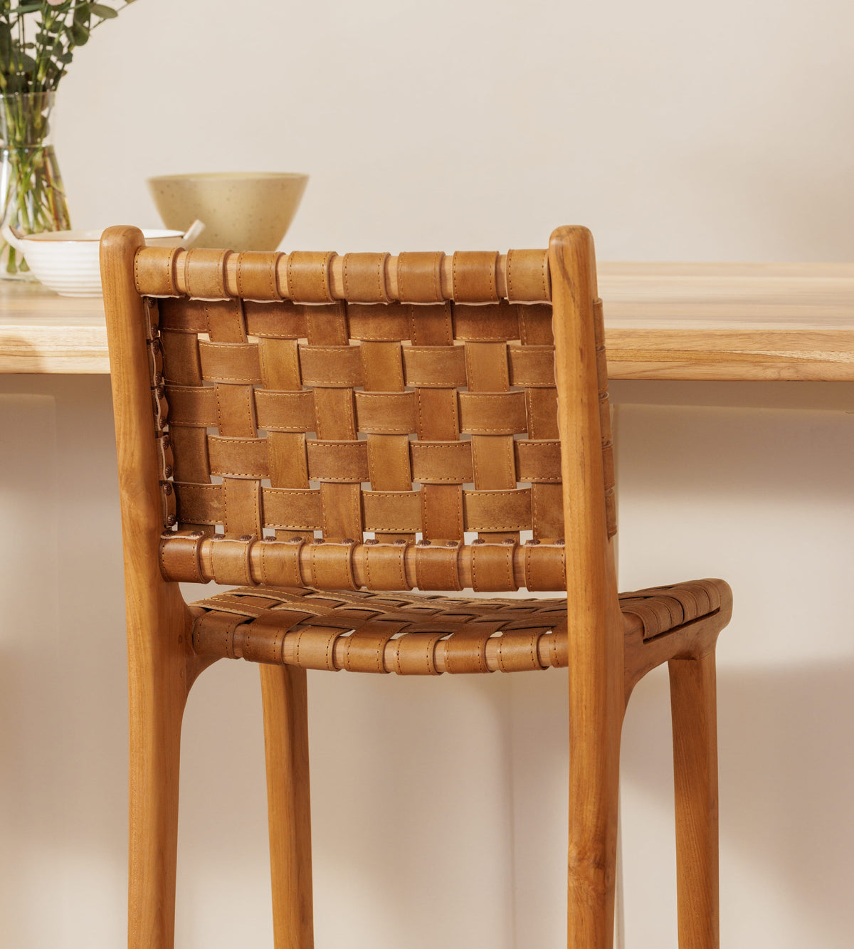 A wooden bar chair with a woven leather seat and back, shown in a warm, minimalist interior setting with a light wood table and neutral background.