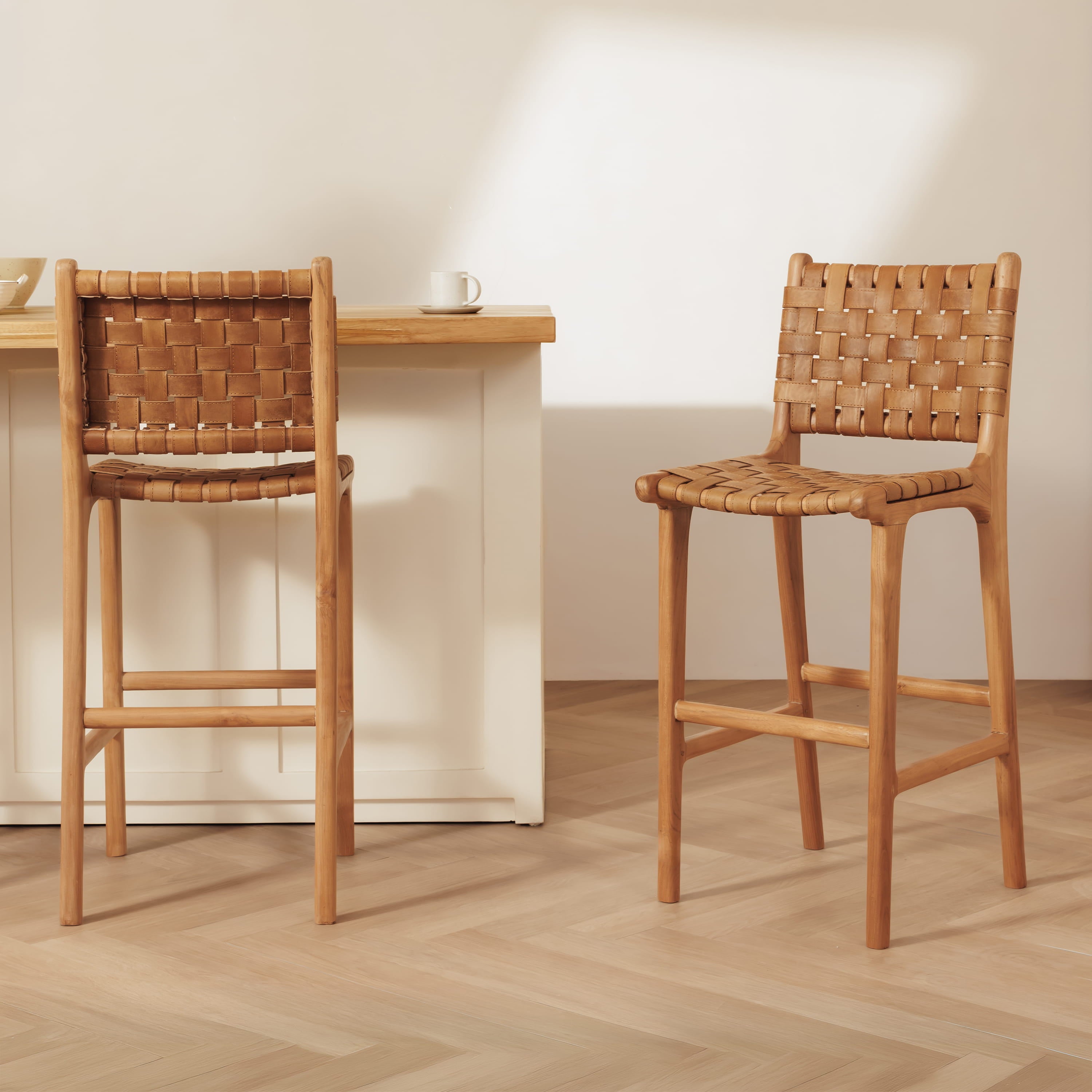 Two wooden bar stools with woven seats and backs, shown in a bright, modern kitchen setting with a light wood countertop and herringbone wood floor.