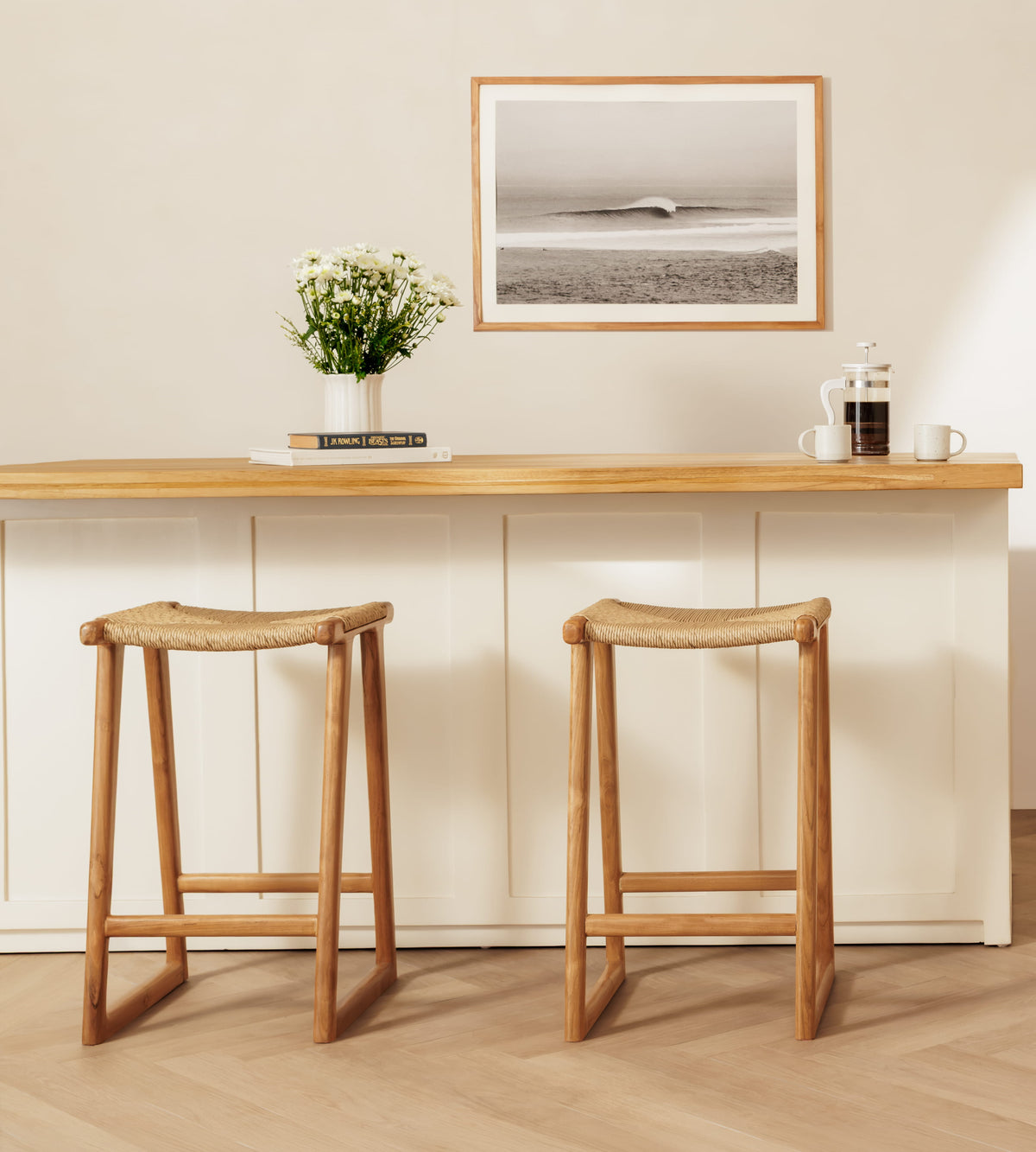 Two wooden bar stools with woven rattan seats are placed in front of a light wood and white counter in a bright, modern interior. A framed black-and-white ocean print hangs on the wall above, with a vase of flowers and small tabletop items on the counter.