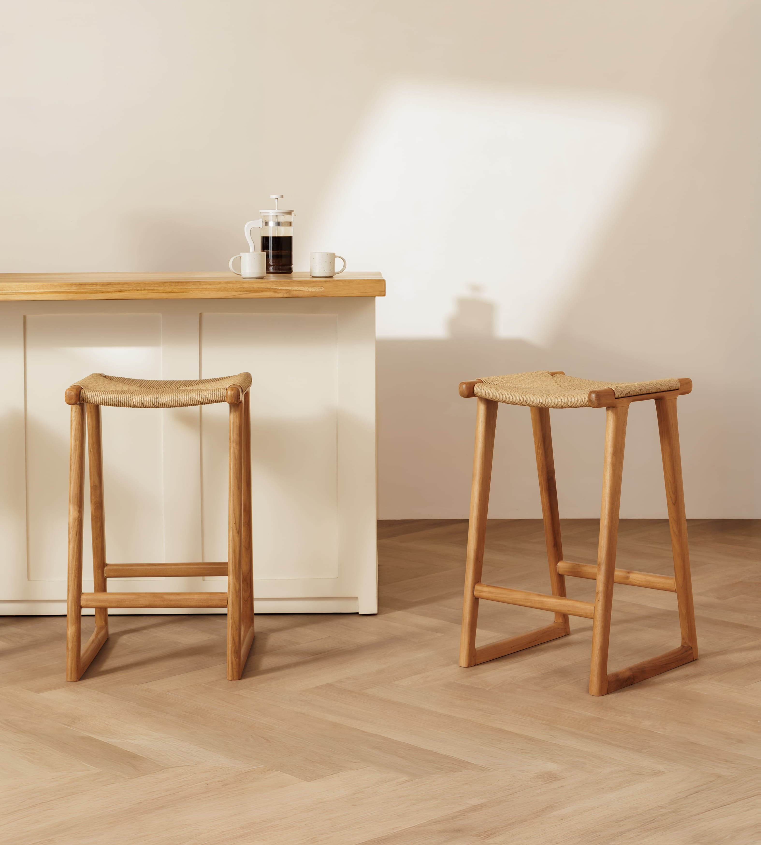 Two wooden backless bar stools with woven rattan seats are shown in a bright, modern interior with a light wood counter and herringbone wood floor.