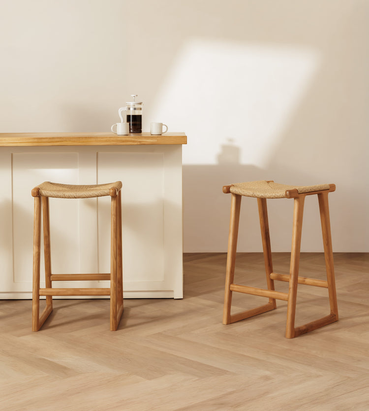 Two wooden backless bar stools with woven rattan seats are shown in a bright, modern interior with a light wood counter and herringbone wood floor.