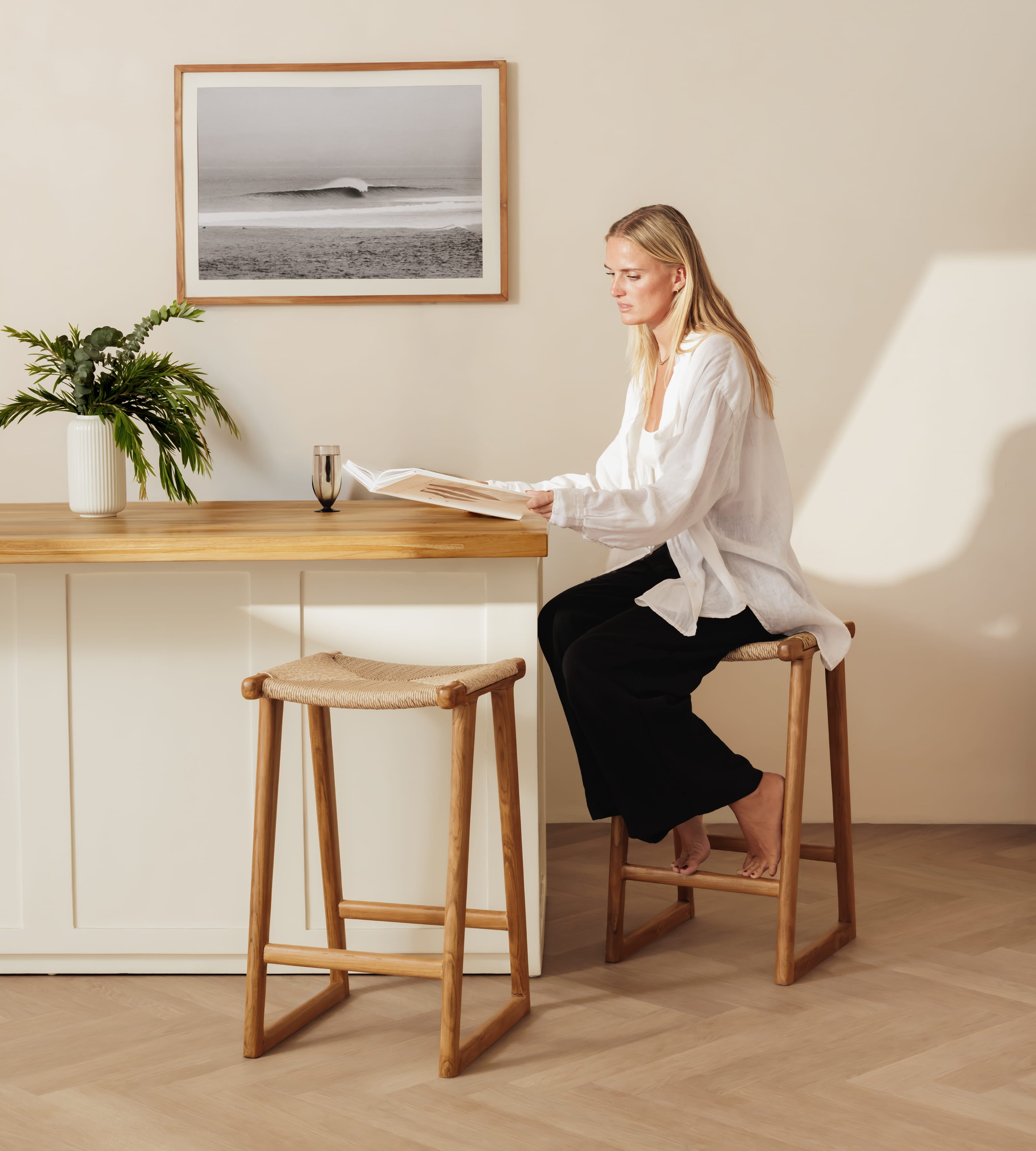 A woman sits on a wooden stool at a bar-height counter in a bright, modern room, reading a magazine. Another matching wooden stool is empty in the foreground.