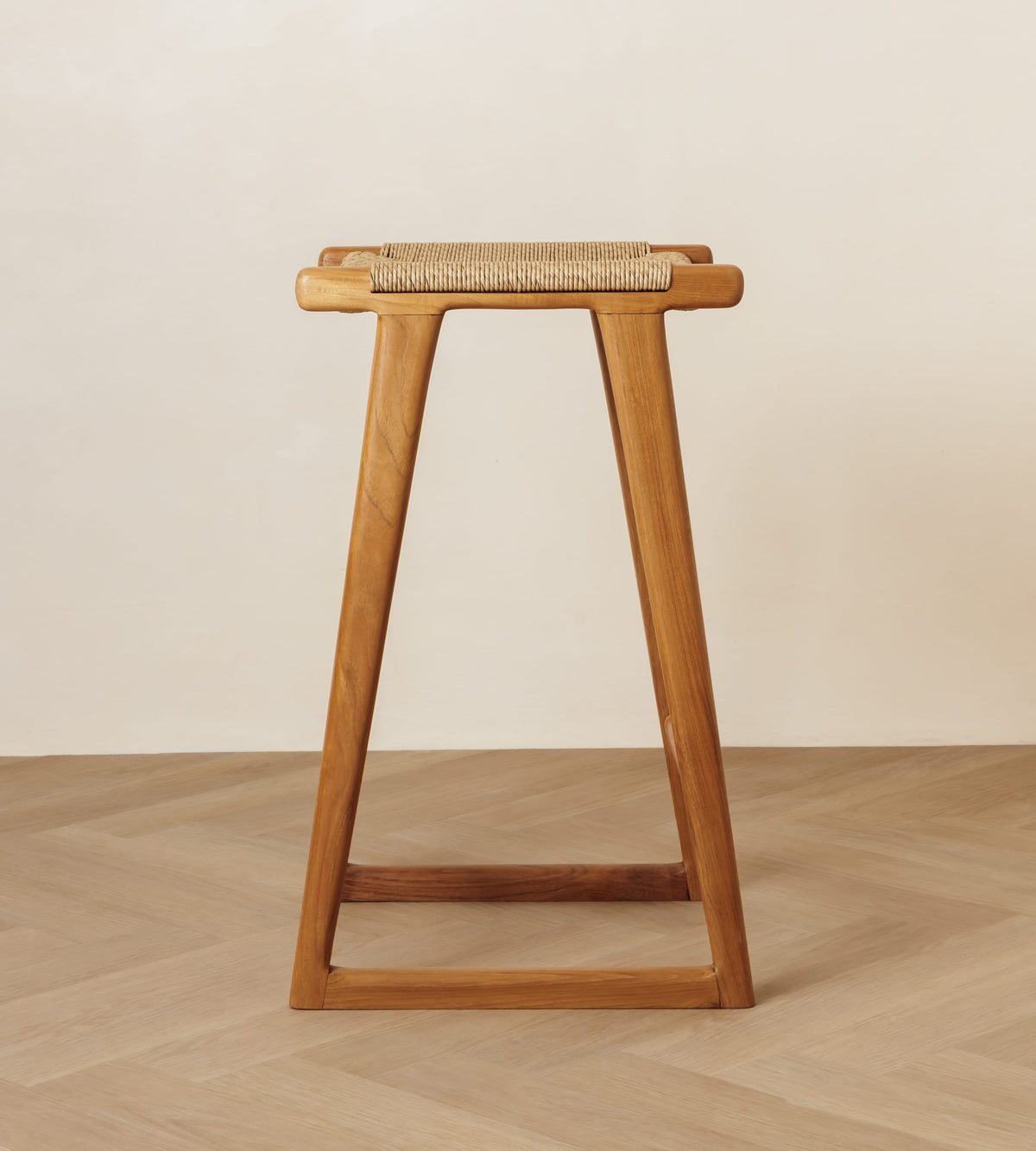 A mid-century modern wooden stool with a woven cane seat, shown from the front against a minimalist beige wall and light wood herringbone floor.