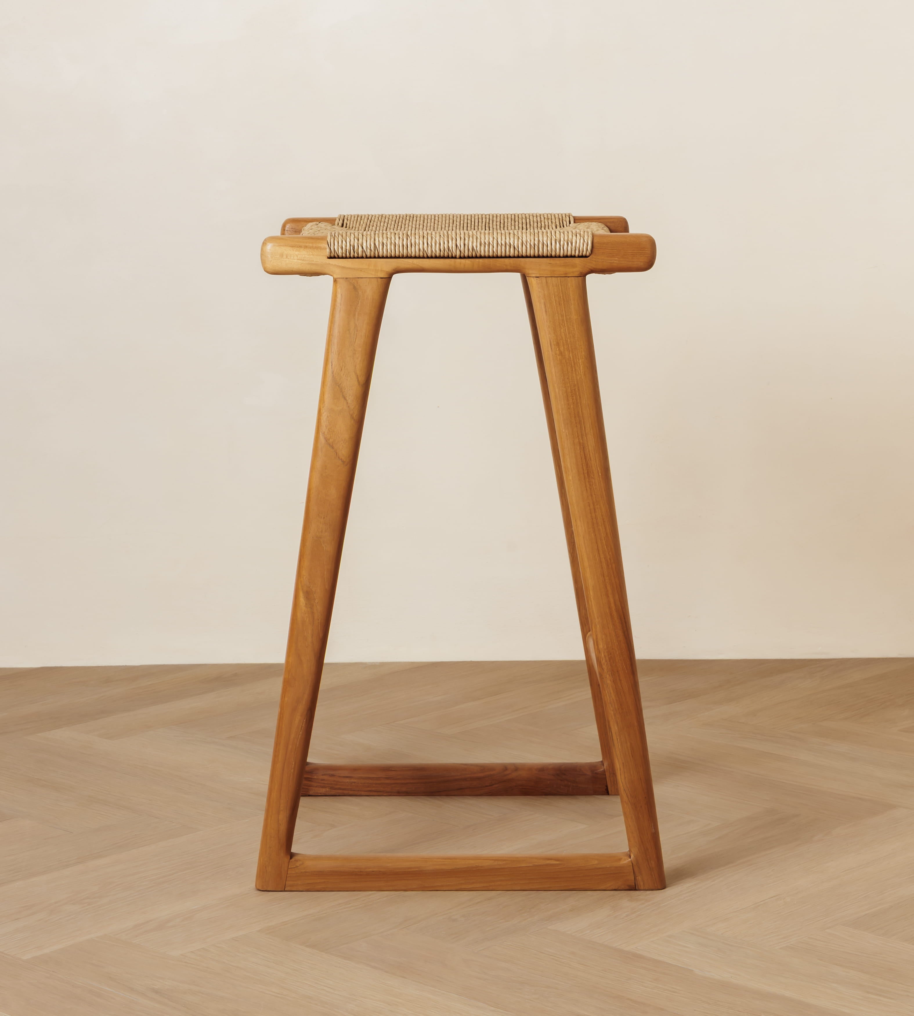 A mid-century modern wooden stool with a woven cane seat, shown from the front against a minimalist beige wall and light wood herringbone floor.