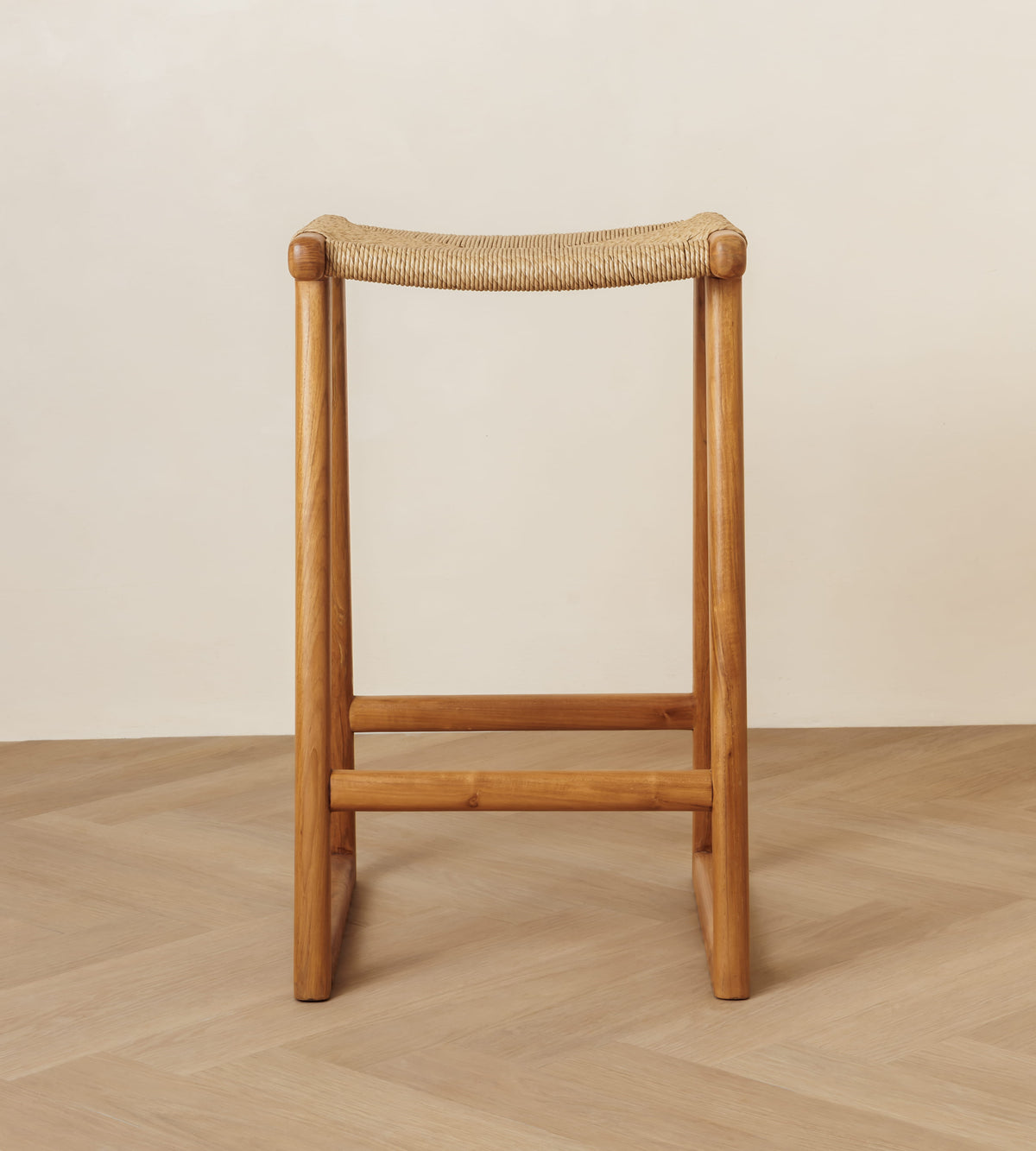 A wooden stool with a woven rope seat, shown from the front against a minimalist beige wall and light wood herringbone floor.