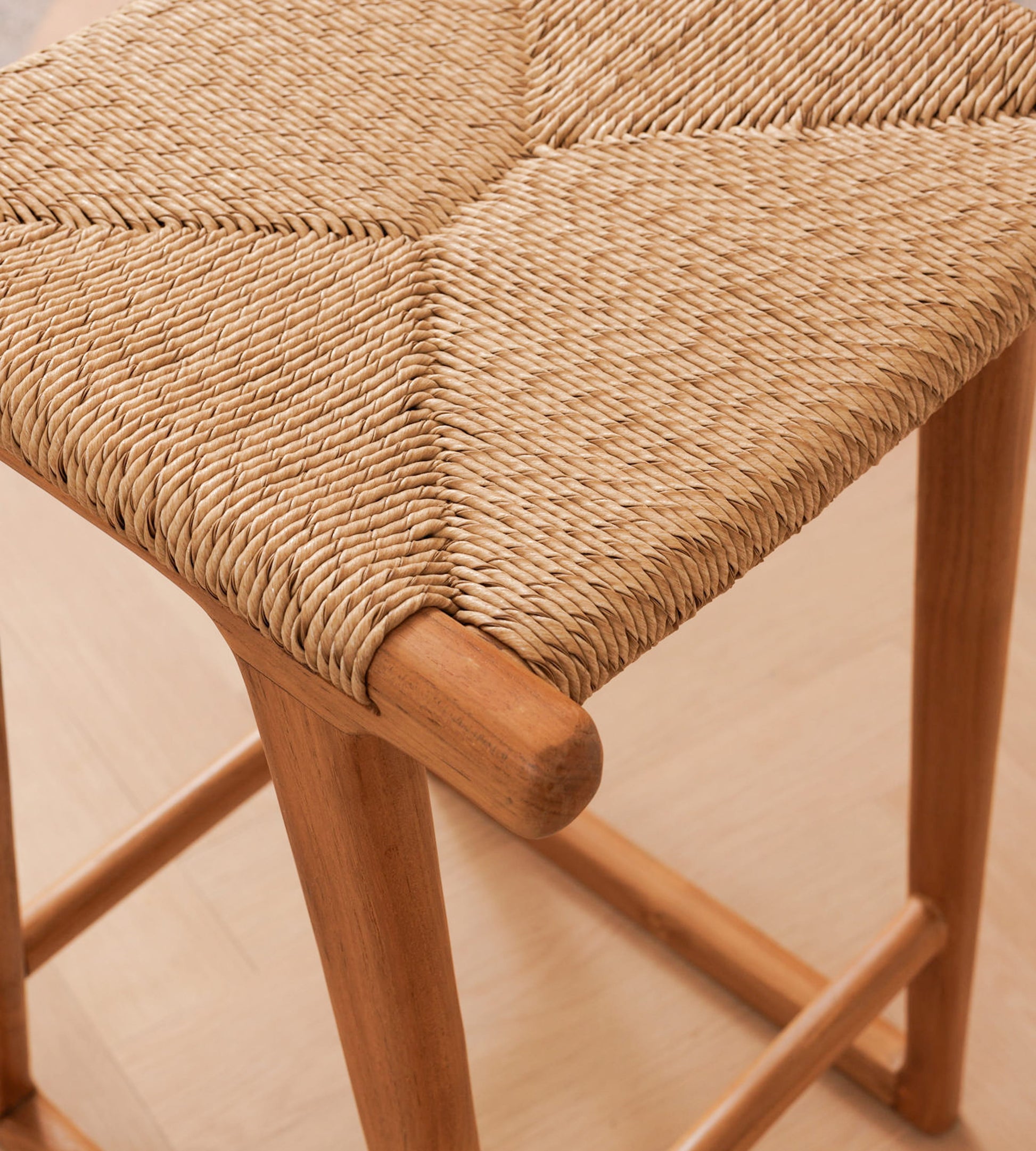 Close-up of a woven rattan seat on a wooden stool, showing the natural tan fiber texture and light wood frame on a pale floor.