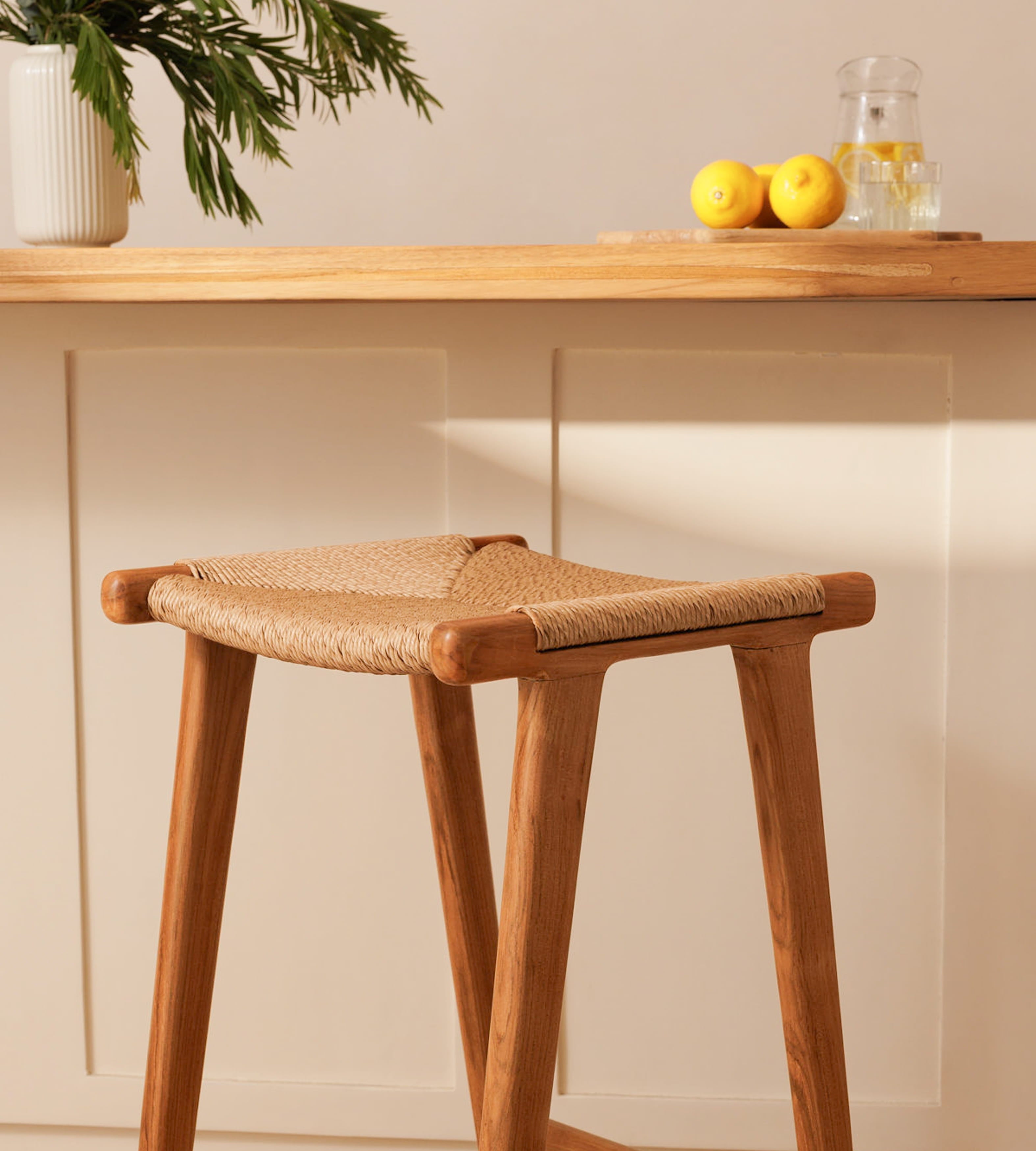 A wooden bar stool with a woven rattan seat cushion, shown in a warm, minimalist kitchen or bar setting with a wooden counter, lemon slices, and a potted plant in the background.