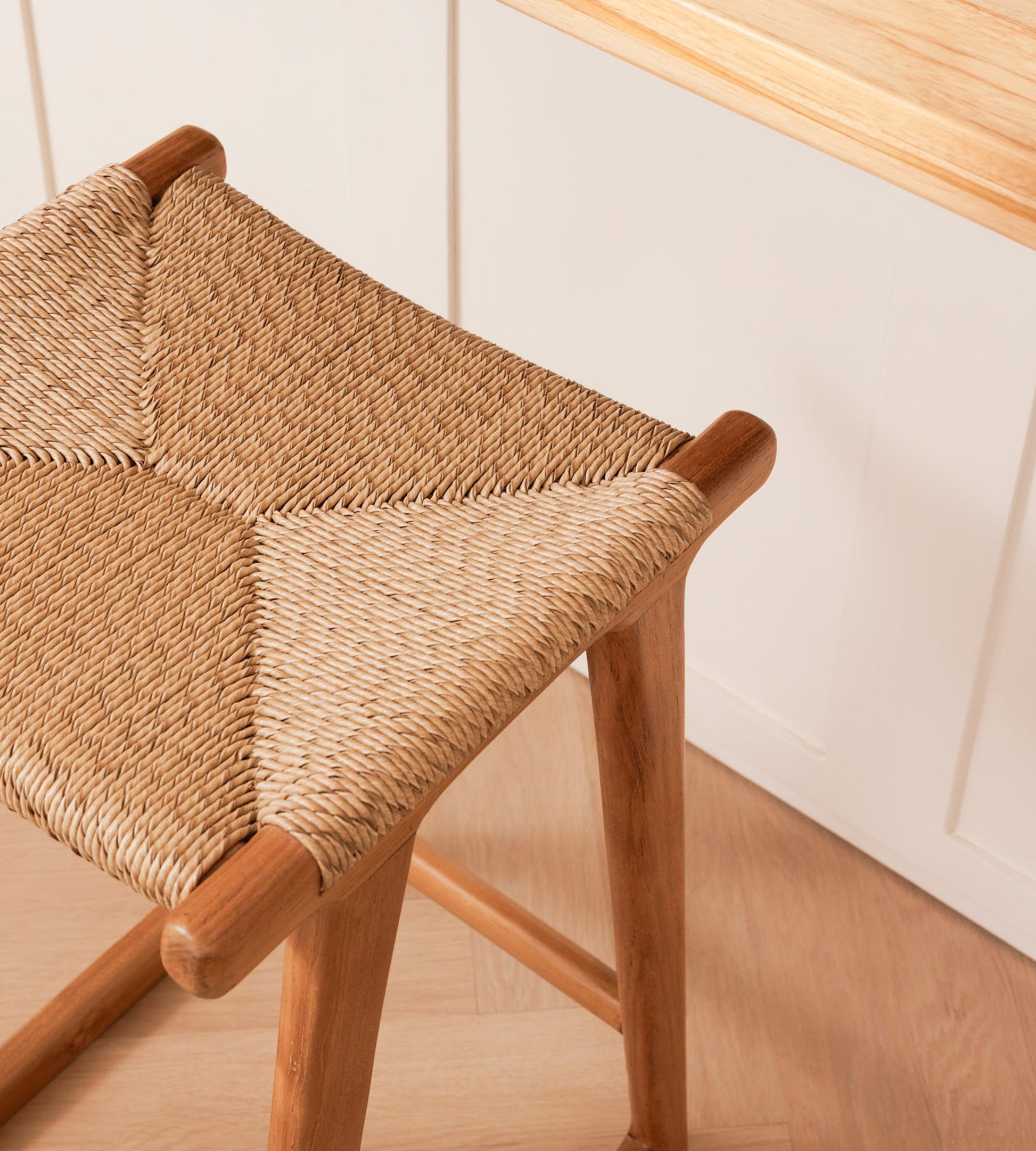 A close-up of a wooden stool with a woven rattan seat, shown in a bright kitchen setting with white cabinets and a light wood countertop.