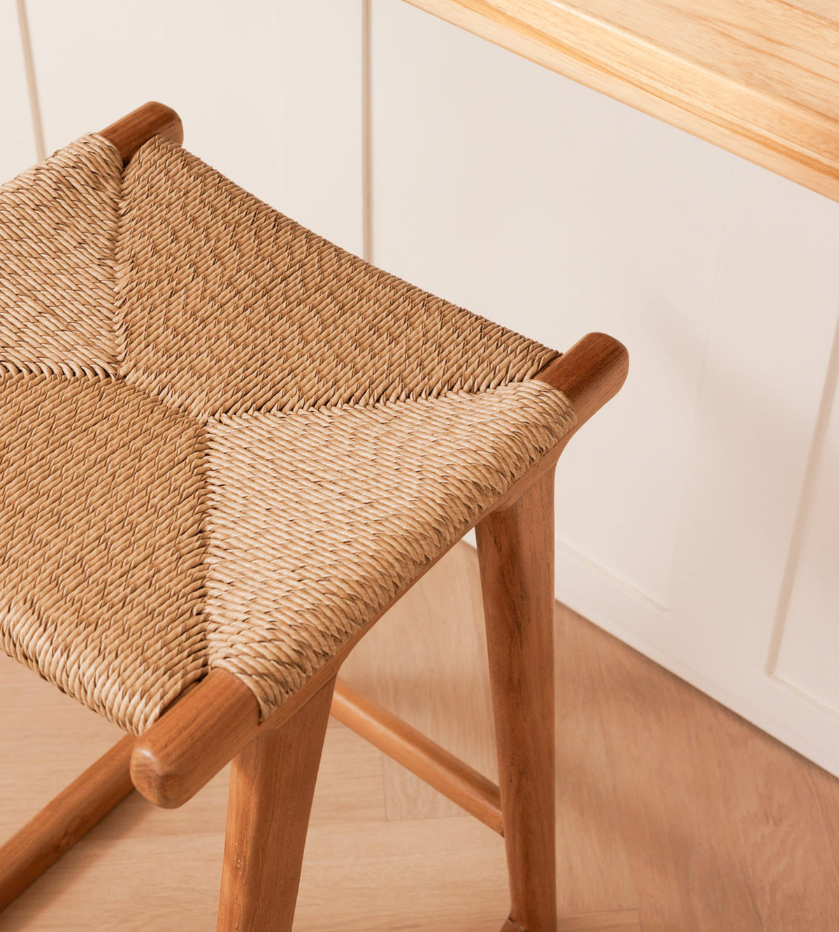 A close-up of a wooden stool with a woven rattan seat, shown in a bright kitchen setting with white cabinets and a light wood countertop.