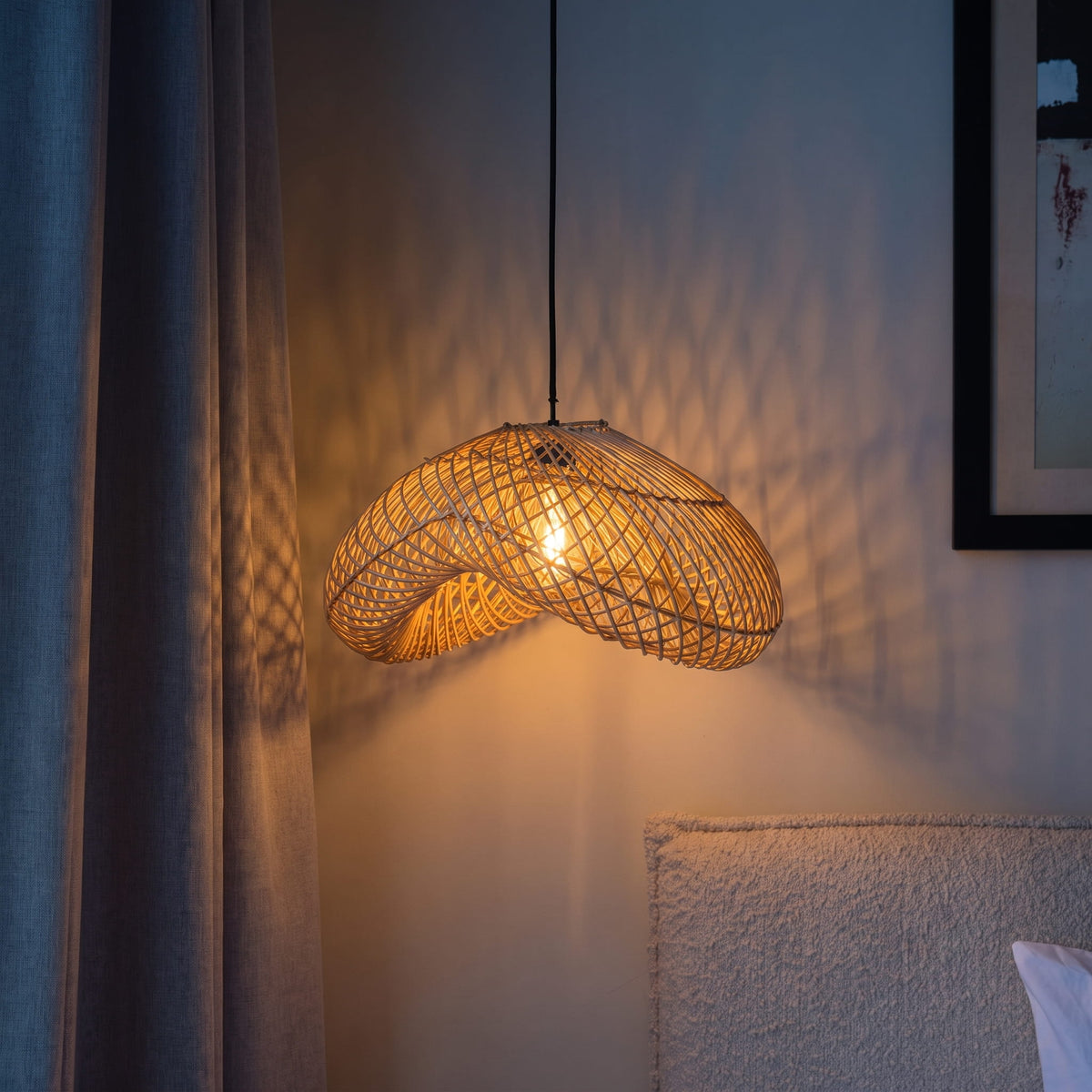 A woven rattan pendant light glowing with a warm yellow light, hanging in a bedroom setting next to a gray curtain and textured wall.