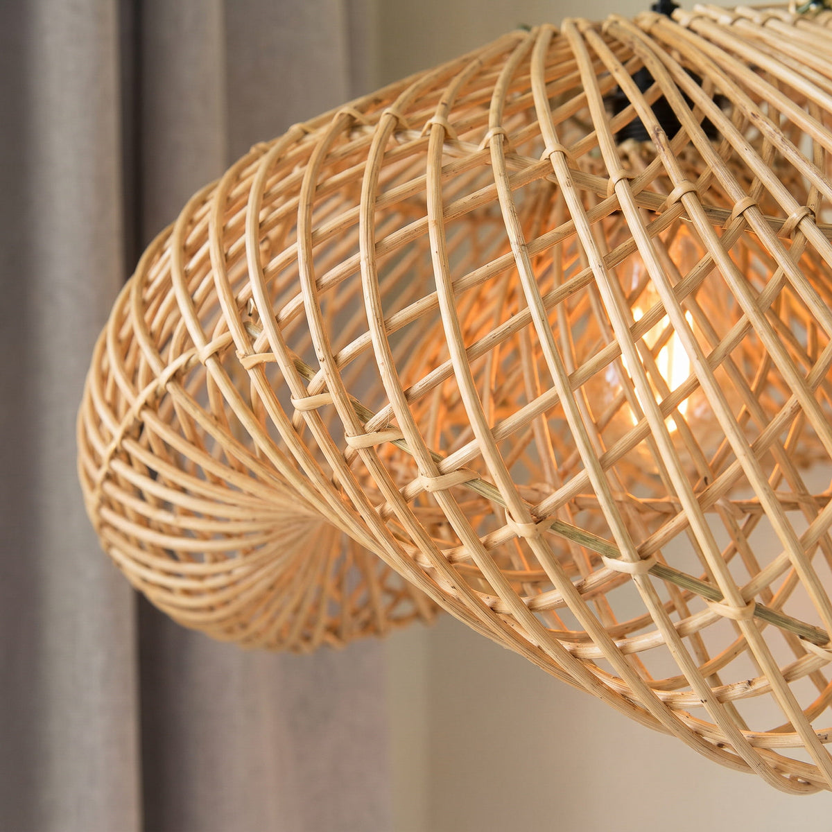 A close-up of a woven natural rattan pendant light shade, showing the detailed hand-woven texture and a warm glowing light bulb inside, set against a soft-focus background of curtains.