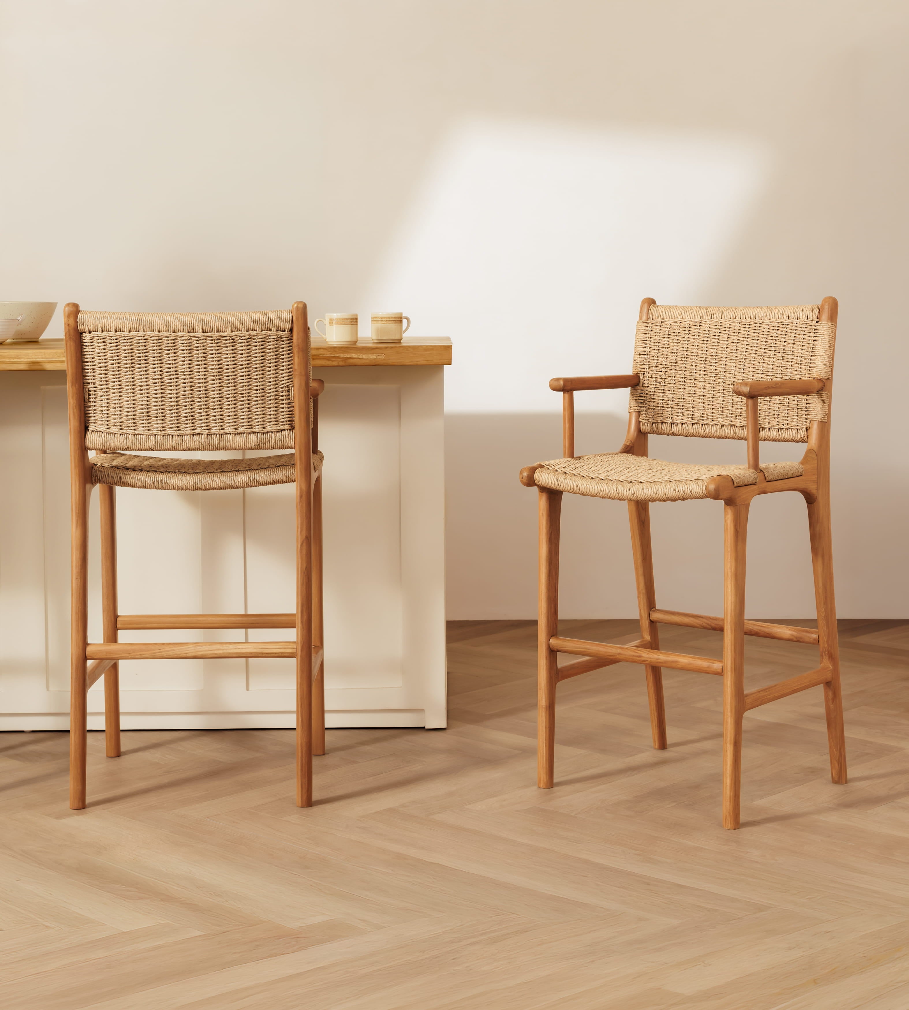Two wooden bar stools with woven rattan seats and backs, shown in a bright minimalist interior with a light wood floor and cream wall.