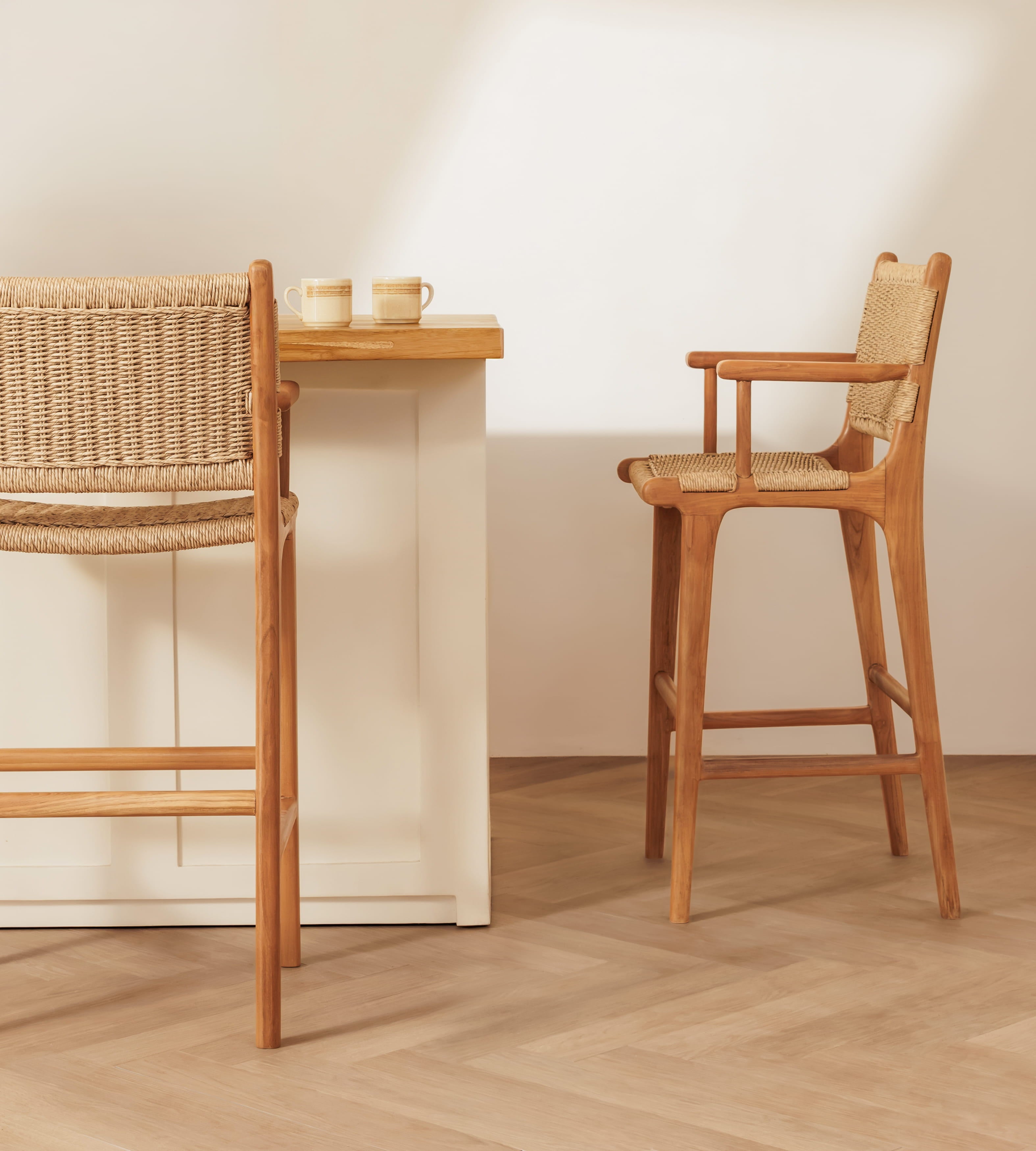 A wooden bar stool with woven cane seats and backrests shown beside a matching wooden counter in a bright, minimalist interior with light wood flooring.