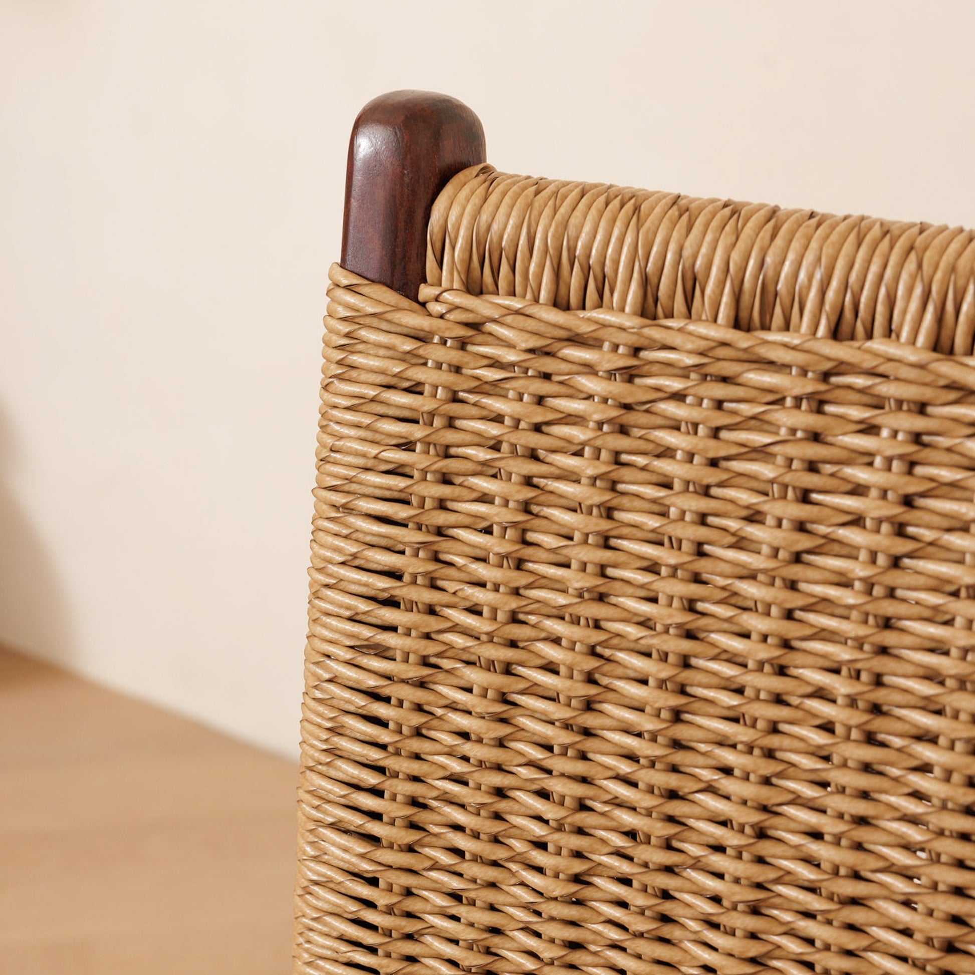 Close-up of a woven rattan or rattan chair back with a dark brown wooden finial on the corner, set against a plain beige wall and light wood floor.