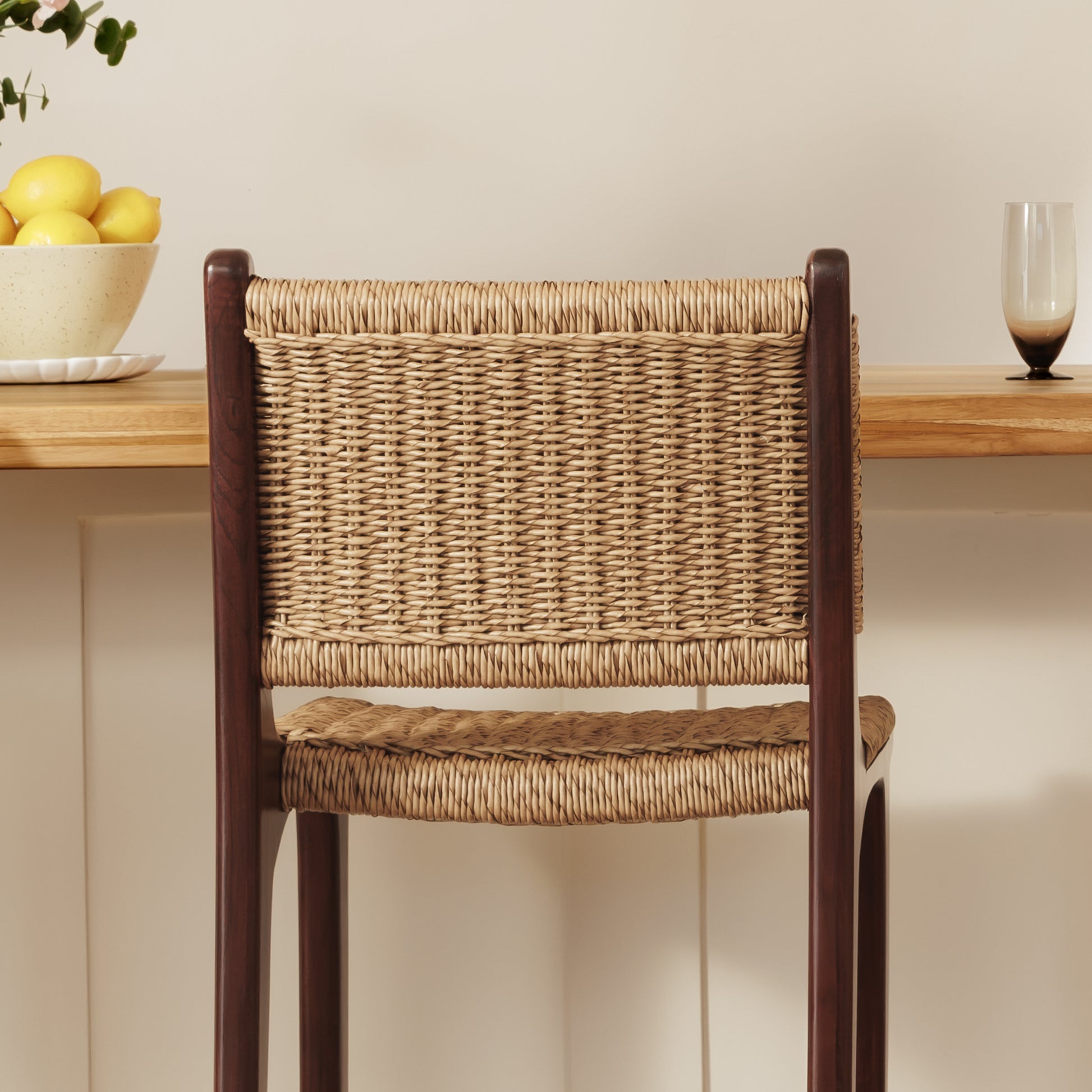 A wooden bar stool with a woven natural-fiber seat and back, shown in a styled interior setting with a wooden shelf, lemons, and a glass in the background.