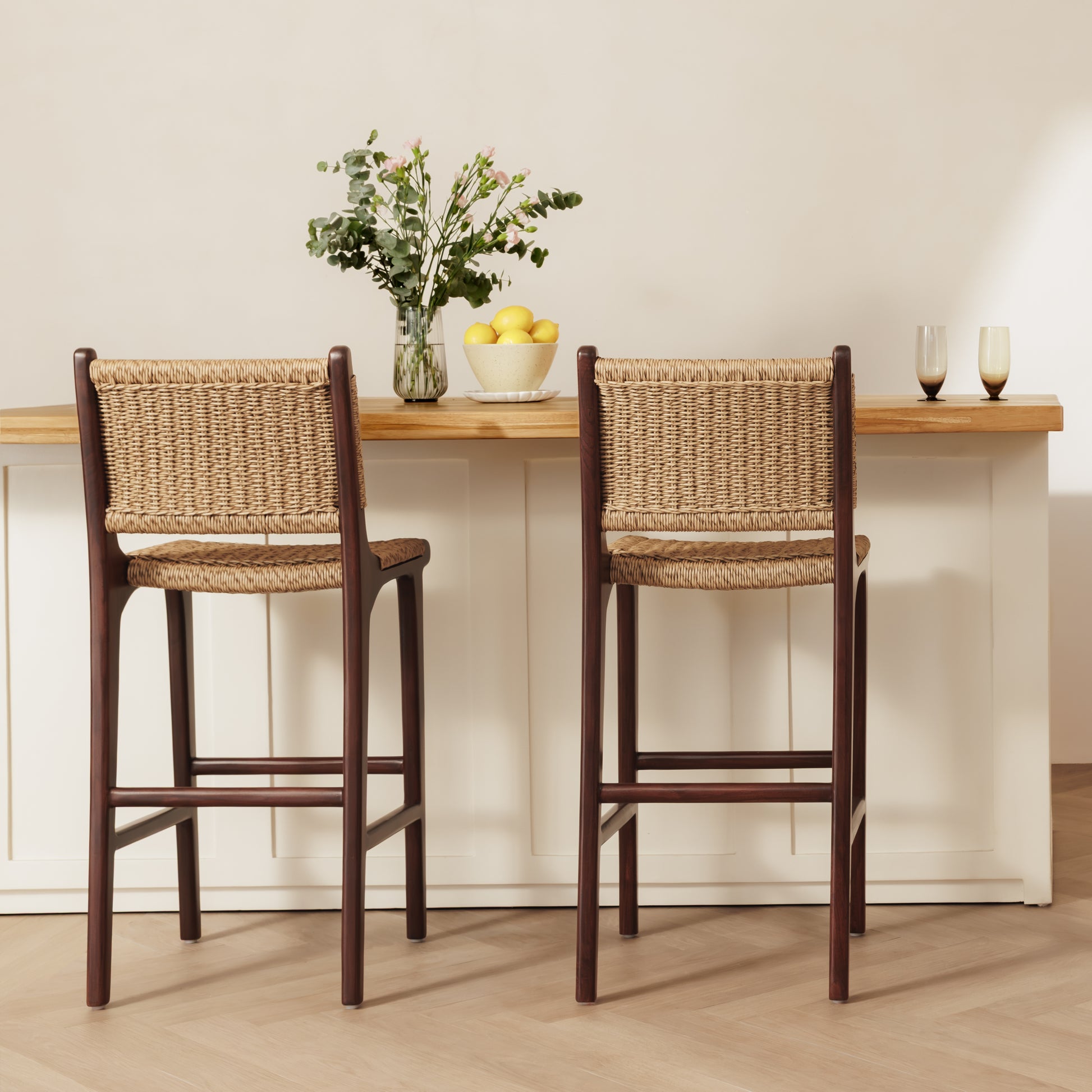 Two rattan bar stools with woven natural-fiber seats and backs, shown in front of a white counter with a light wood top in a bright interior setting.