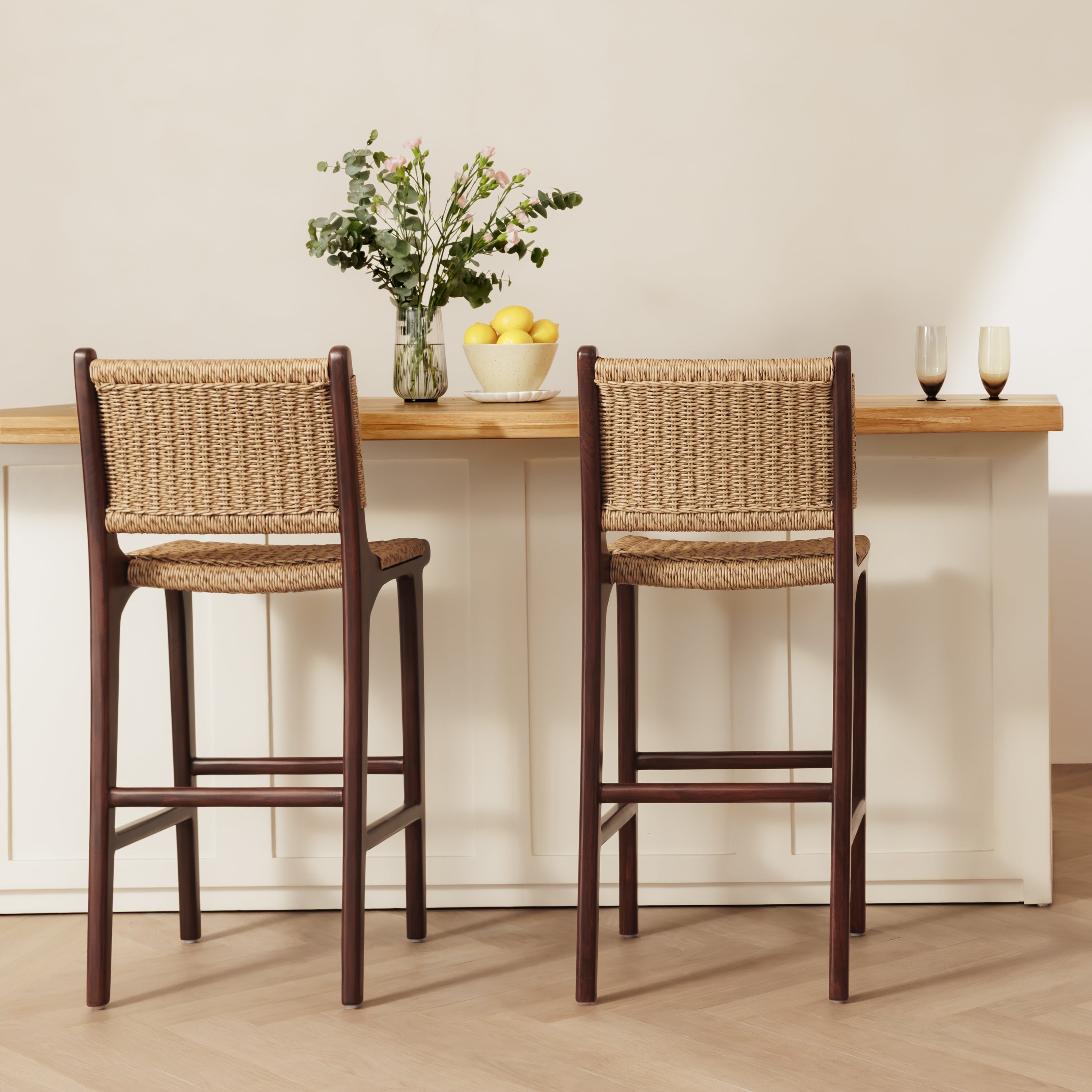 Two rattan bar stools with woven natural-fiber seats and backs, shown in front of a white counter with a light wood top in a bright interior setting.